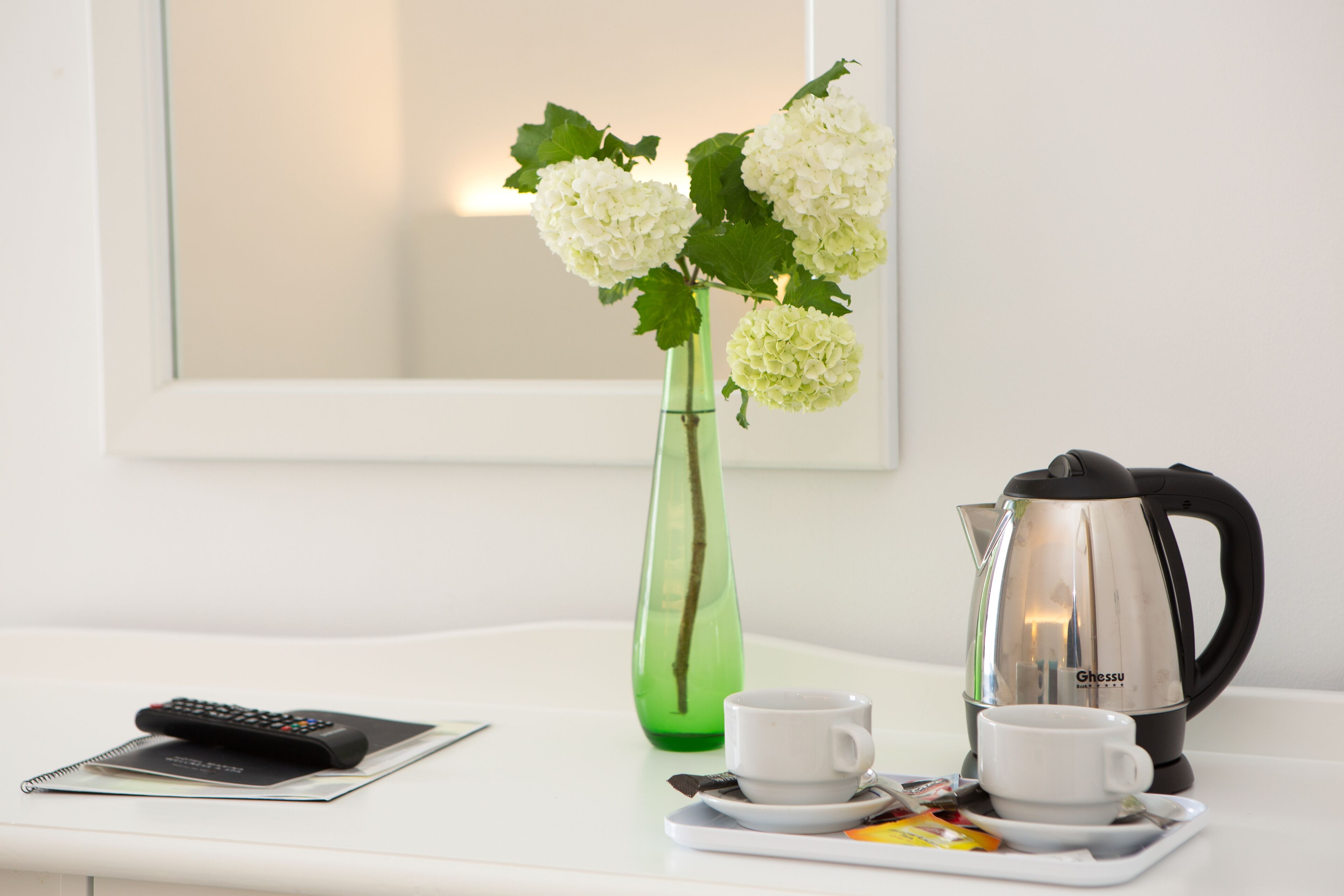 Un plateau avec deux tasses et une théière sur une table blanche, un vase vert avec des fleurs blanches et une télécommande sur un carnet.
