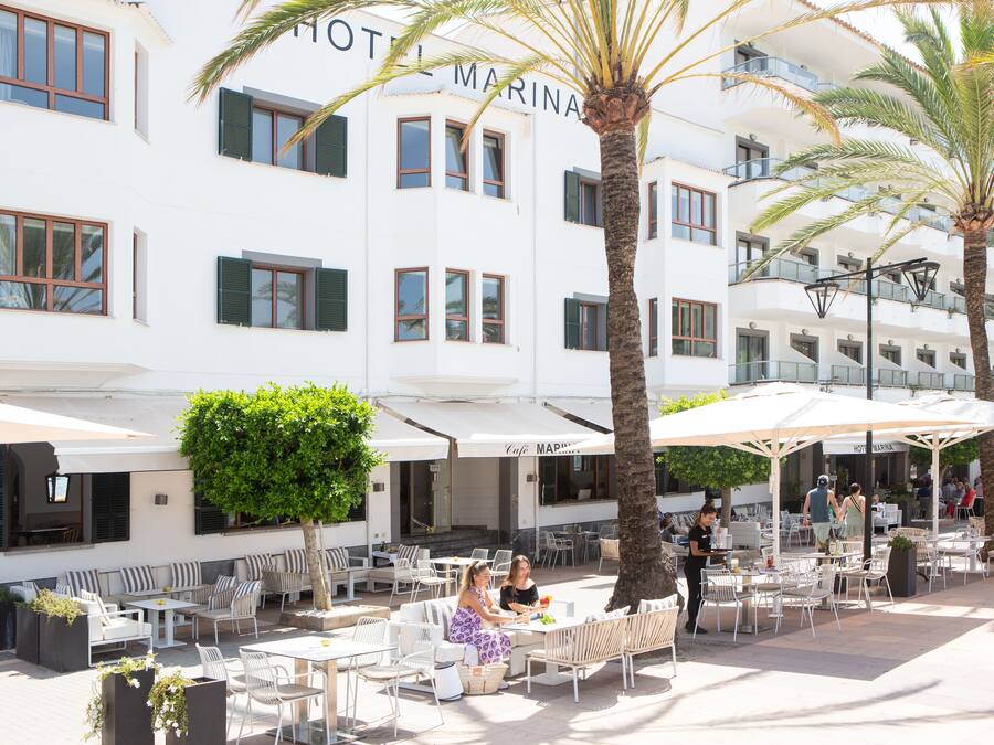 An outdoor seating area with white tables and chairs, palm trees, and a building with a sign reading "HOTEL MARINA".