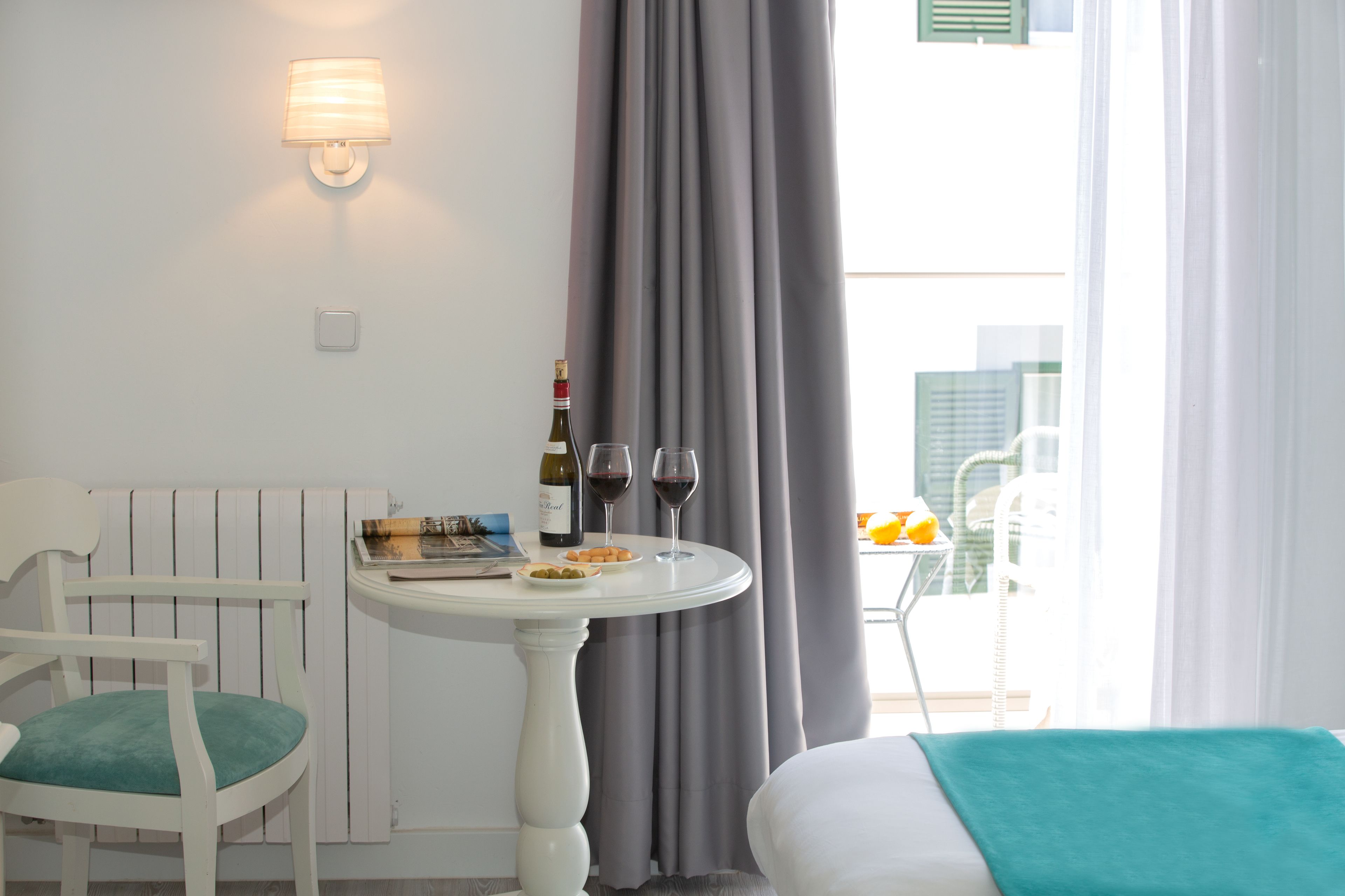 A hotel room with a dining table, a chair, a bed, a window with curtains, and a bottle of wine.