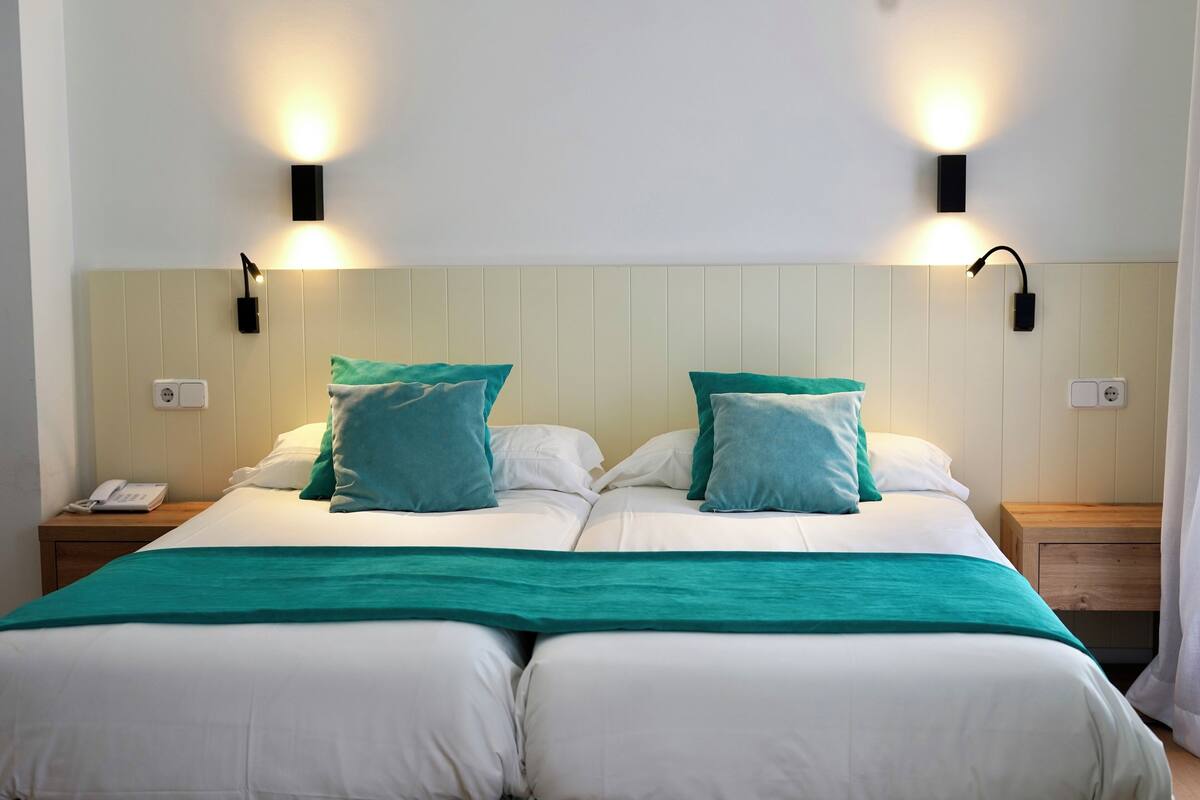 Een tweepersoonsbed met witte en teal beddengoed, twee wandlampen, een bedtelefoon en een houten nachtkastje.