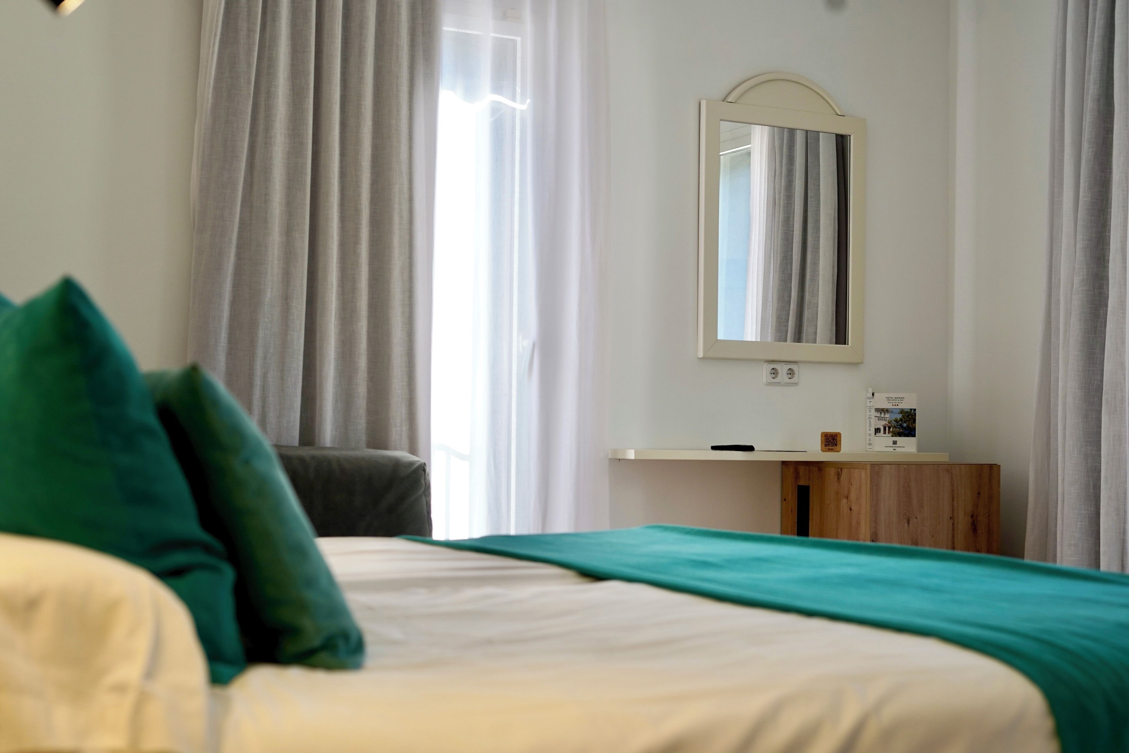 A hotel room with a bed, a bedside table, a mirror, and a window with curtains.
