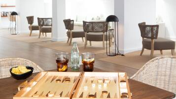Una mesa de madera para juegos de mesa con aperitivos y bebidas, sillas de mimbre y un interior moderno con paredes blancas y suelo de madera.