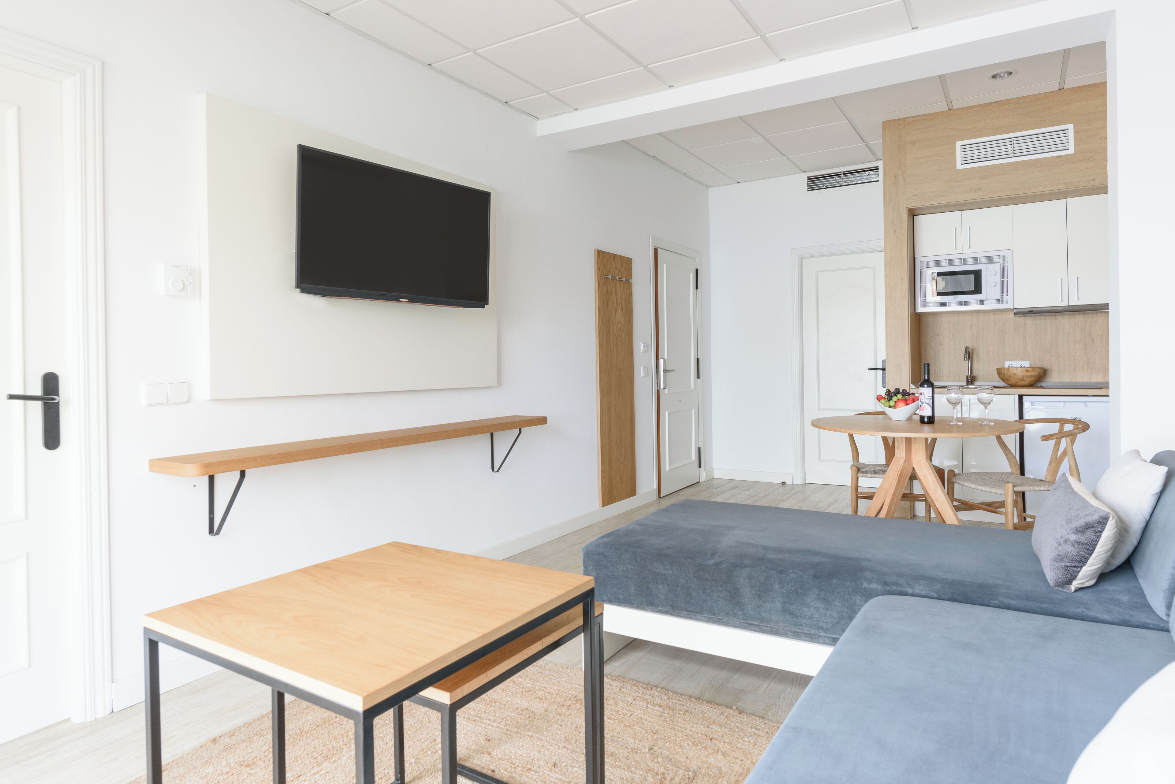 A modern living room with a sofa, a small table, a TV mounted on the wall, and a kitchen area with a microwave.