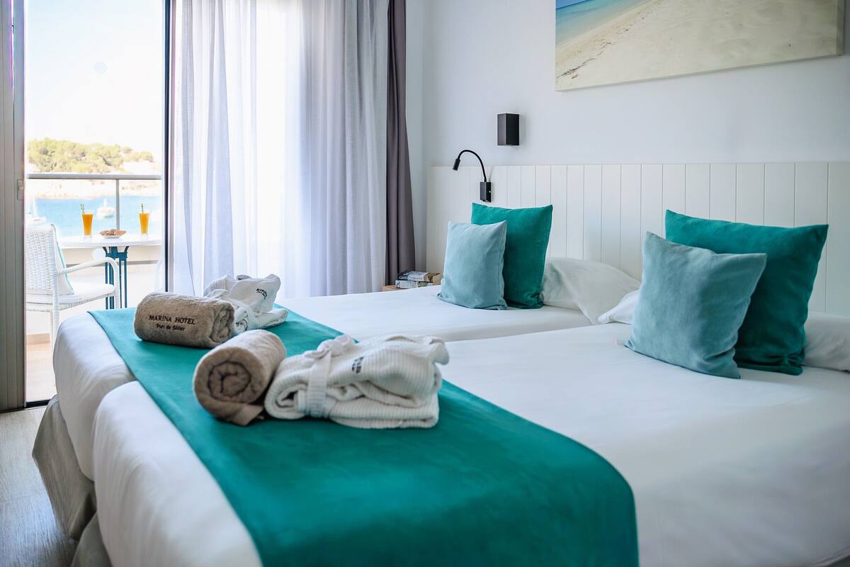 Een hotelkamer met twee bedden, blauwgroene beddengoed, uitzicht op zee, een balkon met een tafel en stoelen, en een schilderij aan de muur.