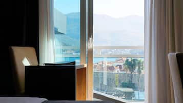 Habitación de hotel con cama, escritorio y vistas a montañas y un paisaje urbano a través de amplios ventanales.