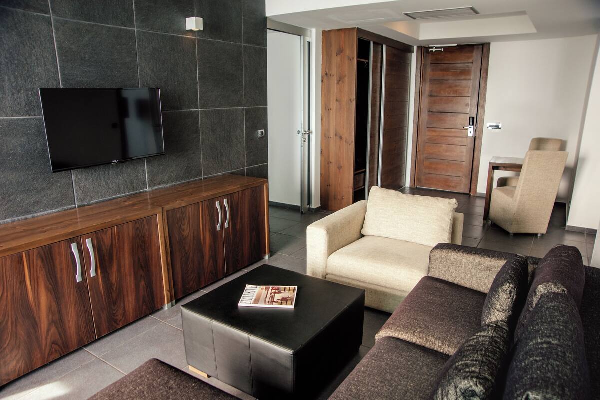A modern living room with a flat-screen TV mounted on a dark tiled wall, a wooden cabinet with silver handles, a black ottoman with a magazine, a beige sofa, and a dark-colored couch.