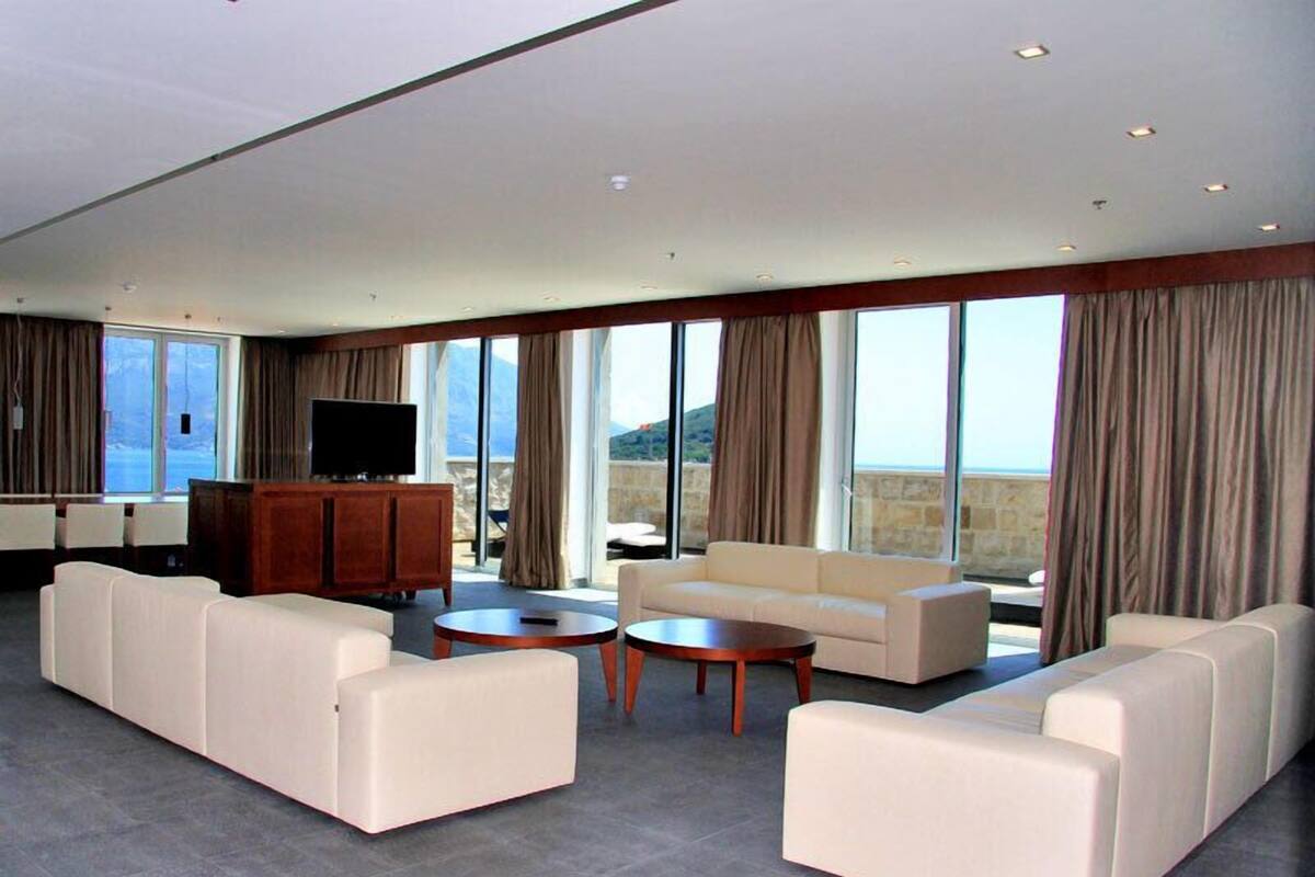 A spacious living room with a large sofa set, a coffee table, and a flat-screen TV. The room has large windows with curtains, offering a view of the outdoors.