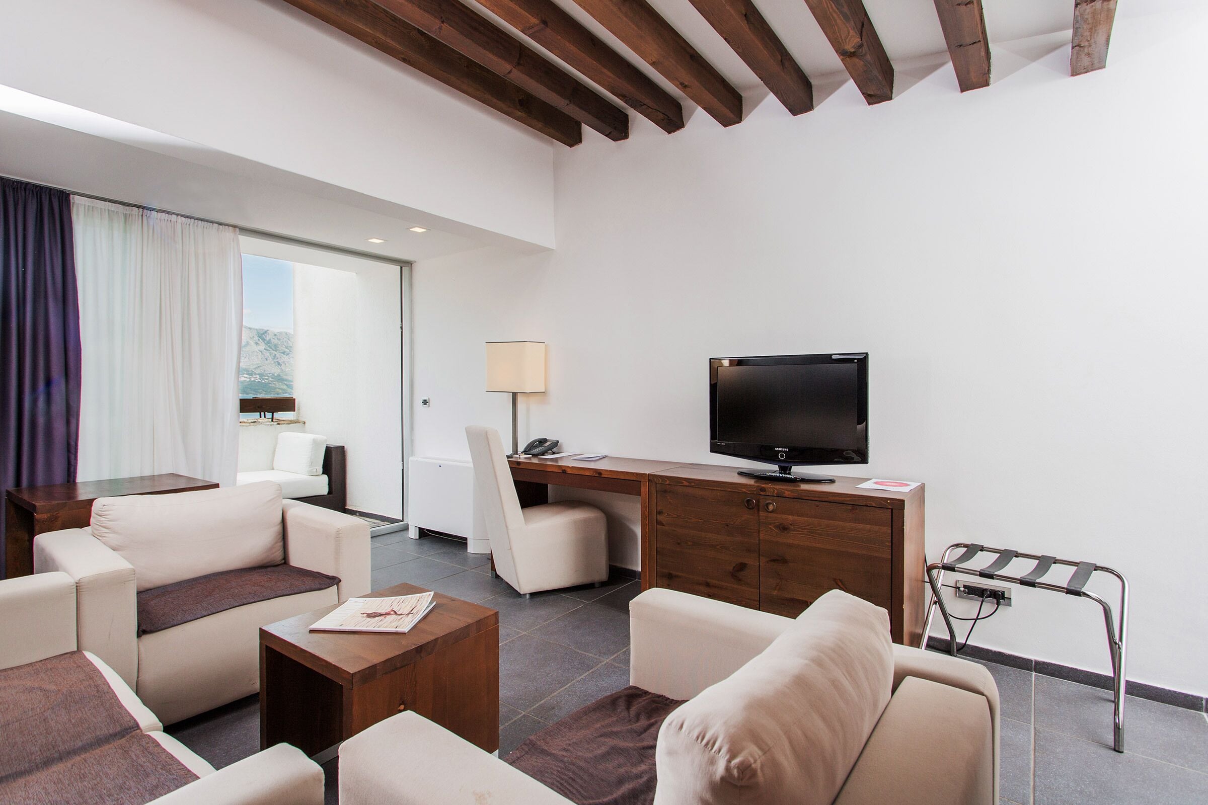 A modern hotel room with a sofa, armchair, coffee table, desk with a flat-screen TV, and a view of mountains through the window.