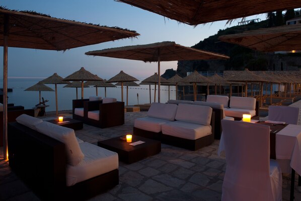 An outdoor seating area with cushioned sofas and tables, surrounded by thatched umbrellas, overlooking a body of water.