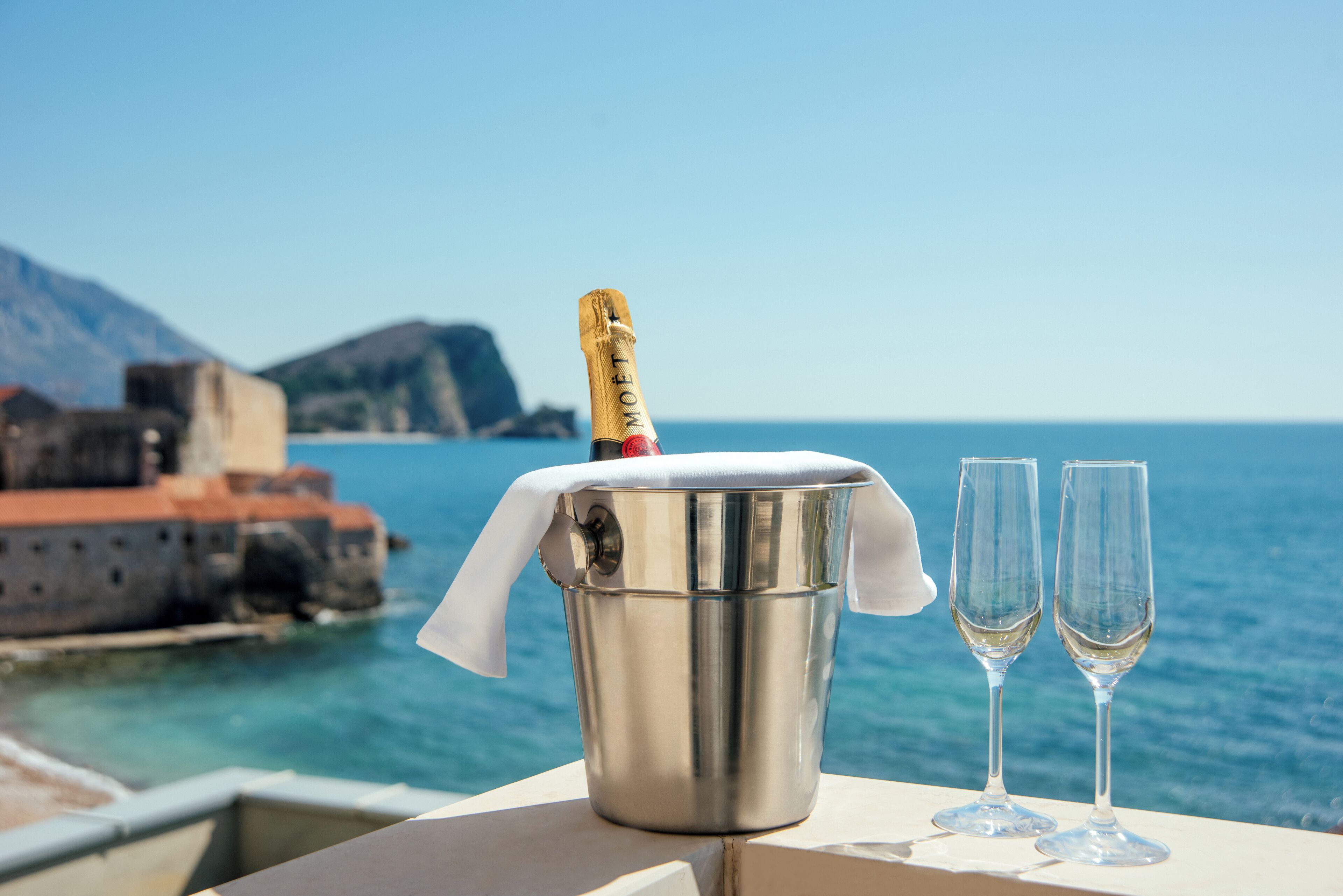 A metallic bucket with a bottle of champagne, two empty glasses, and a white cloth on a table with a coastal view in the background.