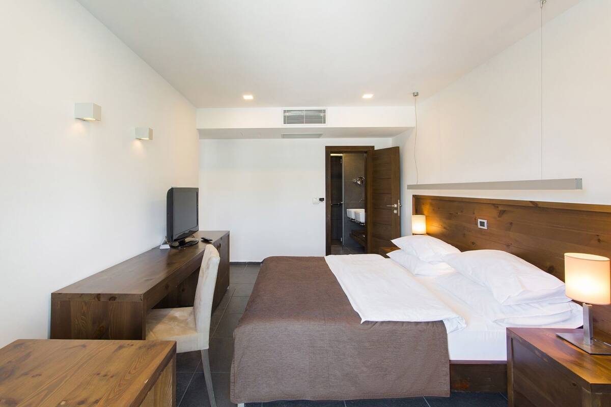 A hotel room with a bed, a desk with a television, a chair, a bedside table with a lamp, and a bathroom door.