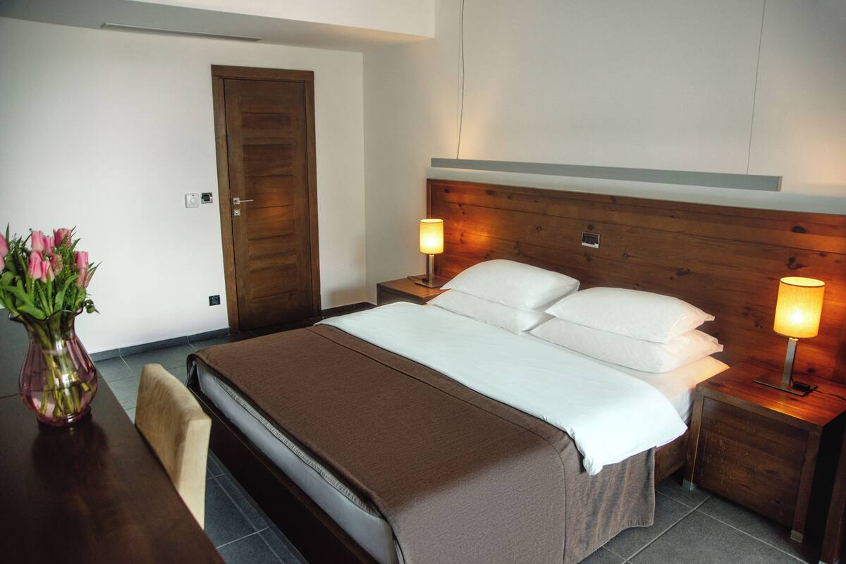 A hotel room with a wooden headboard, a bed with white linens, two bedside tables with lamps, a wooden nightstand, a chair, and a vase with flowers.