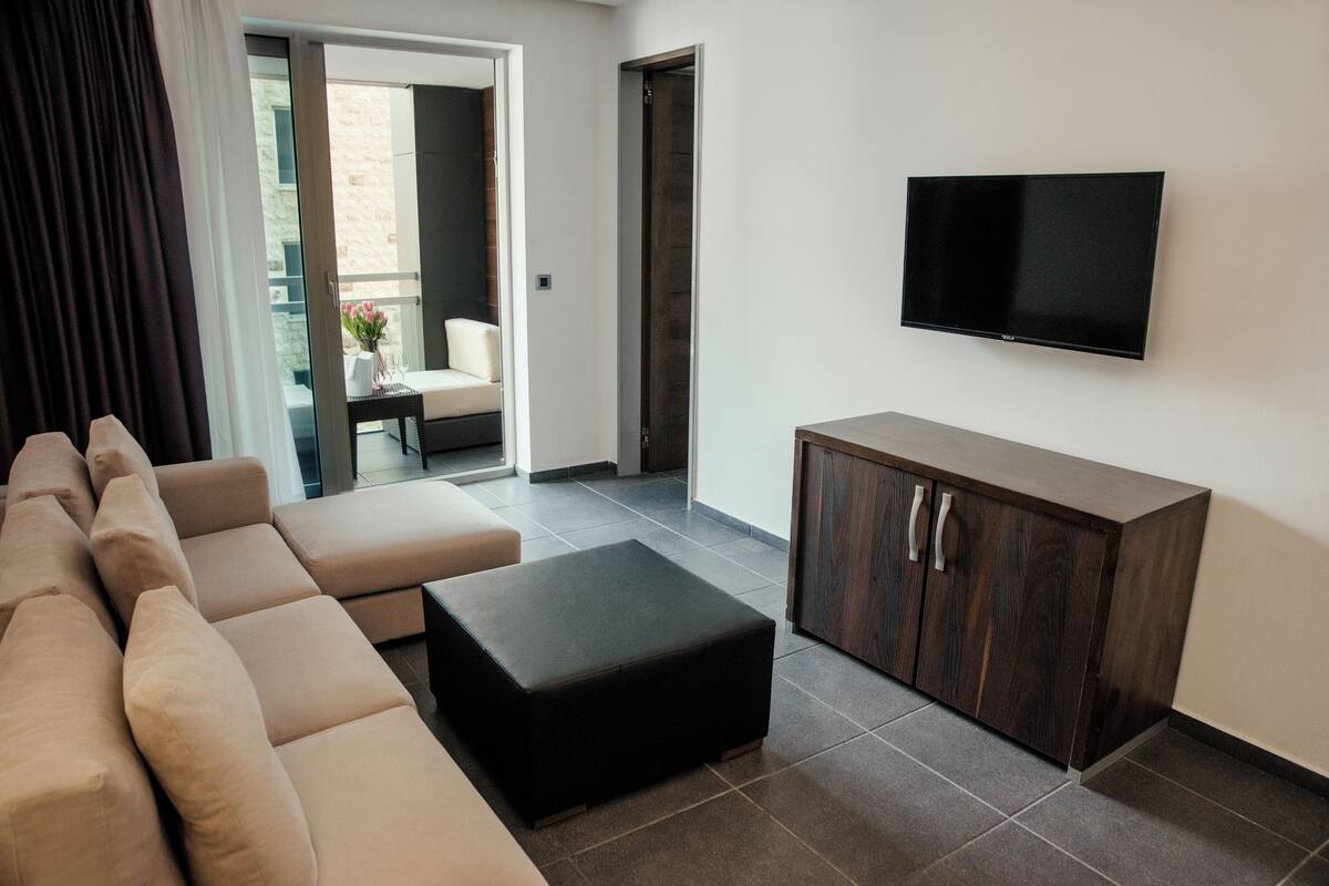 A modern living room with a sectional sofa, a black ottoman, a wooden cabinet, and a flat-screen TV mounted on the wall.