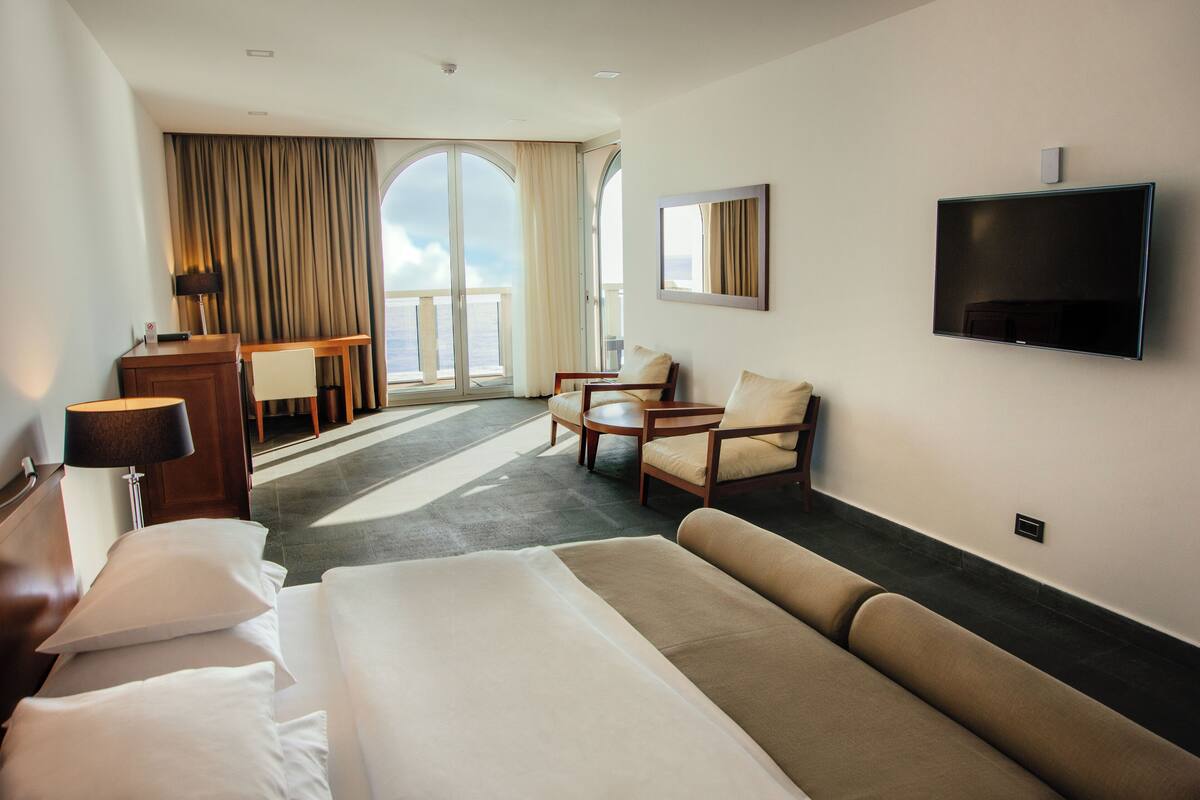 A hotel room with a large bed, a desk, a chair, a sofa, and a TV mounted on the wall.