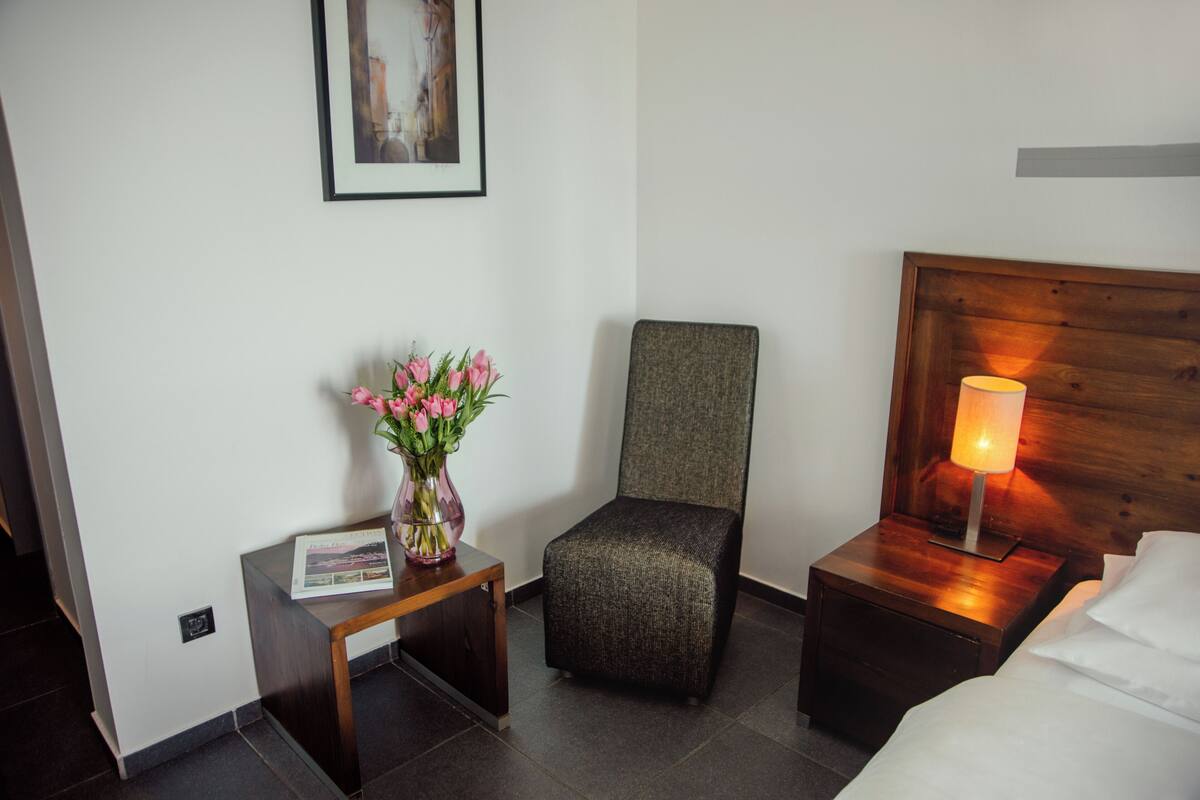 A hotel room with a bed, a nightstand with a lamp, a small table with a vase of flowers, a chair, and a framed picture on the wall.
