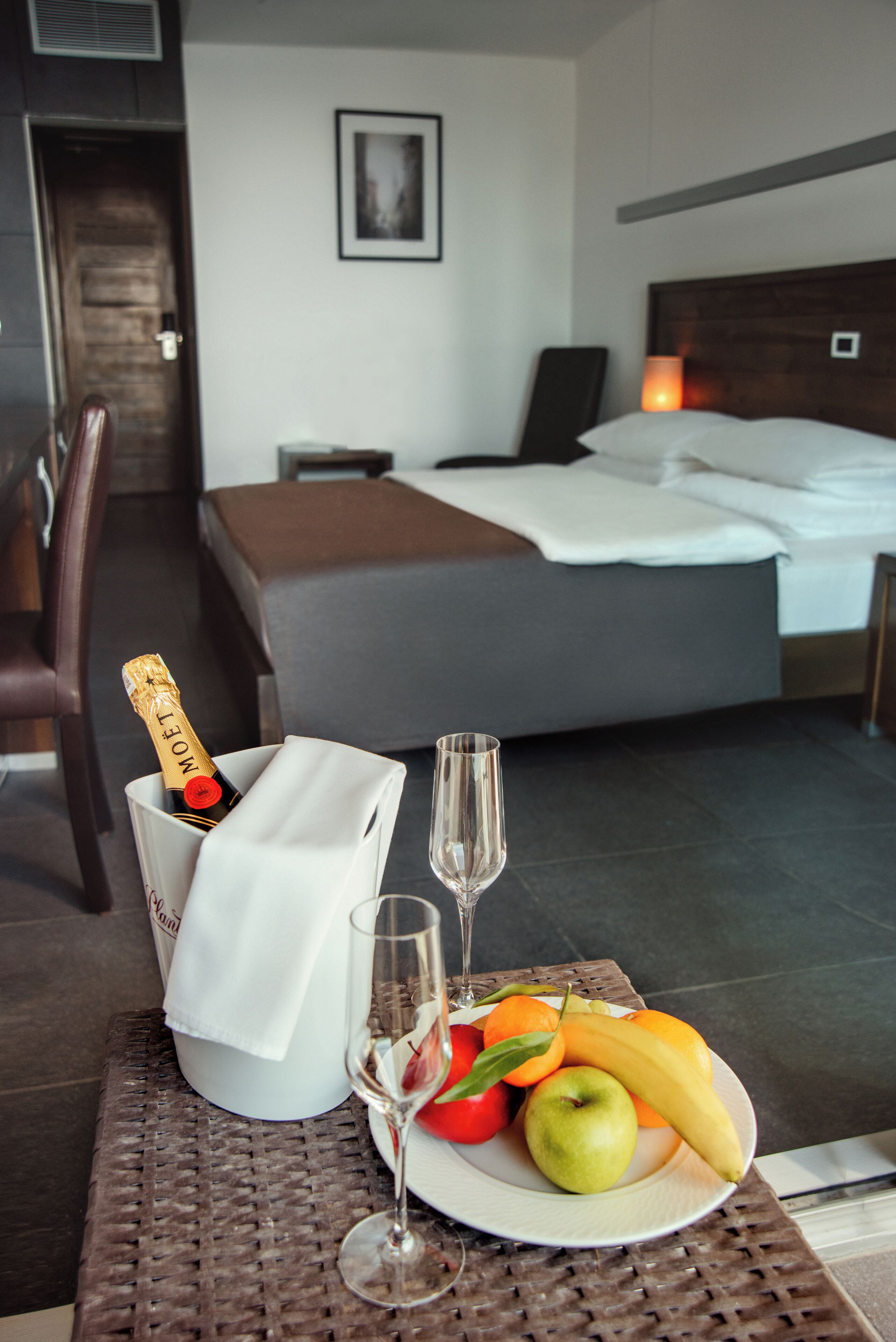 Una habitación de hotel con una cama, una mesita de noche con una lámpara, una botella de champán, una copa y un plato de fruta.