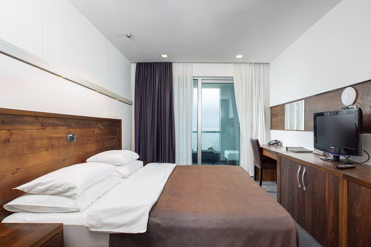 A hotel room with a wooden headboard, a bed with white linens, a wooden desk with a television, and a view of a balcony through a glass door.