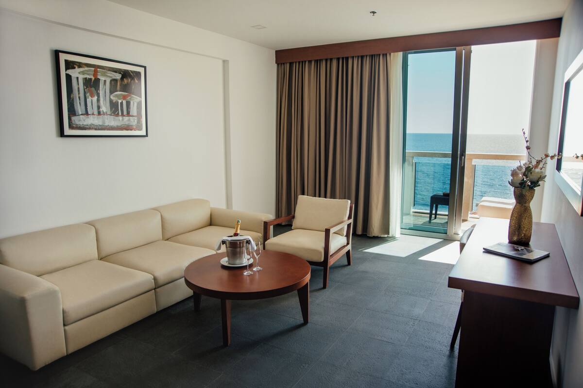 A hotel room with a sofa, armchair, and a small table with a bottle and glasses. A painting is on the wall, and there's a view of the sea through large windows.