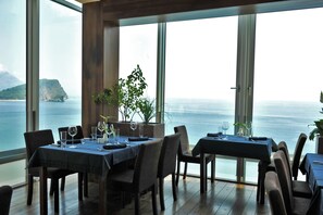 Un espace repas avec des tables préparées pour deux personnes, de grandes fenêtres offrant une vue sur la mer et une île au loin.