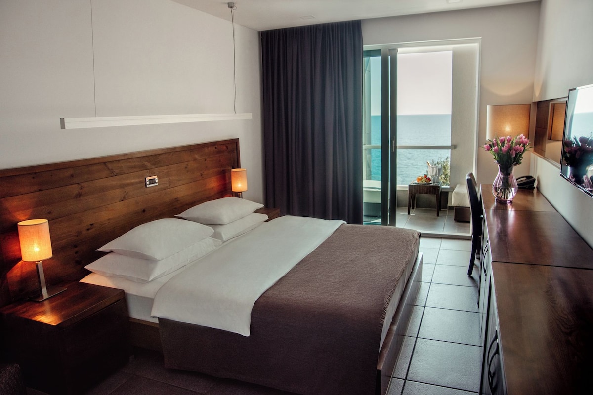 A hotel room with a bed, bedside lamps, a TV, a vase with flowers, and a view of the sea through a sliding door.