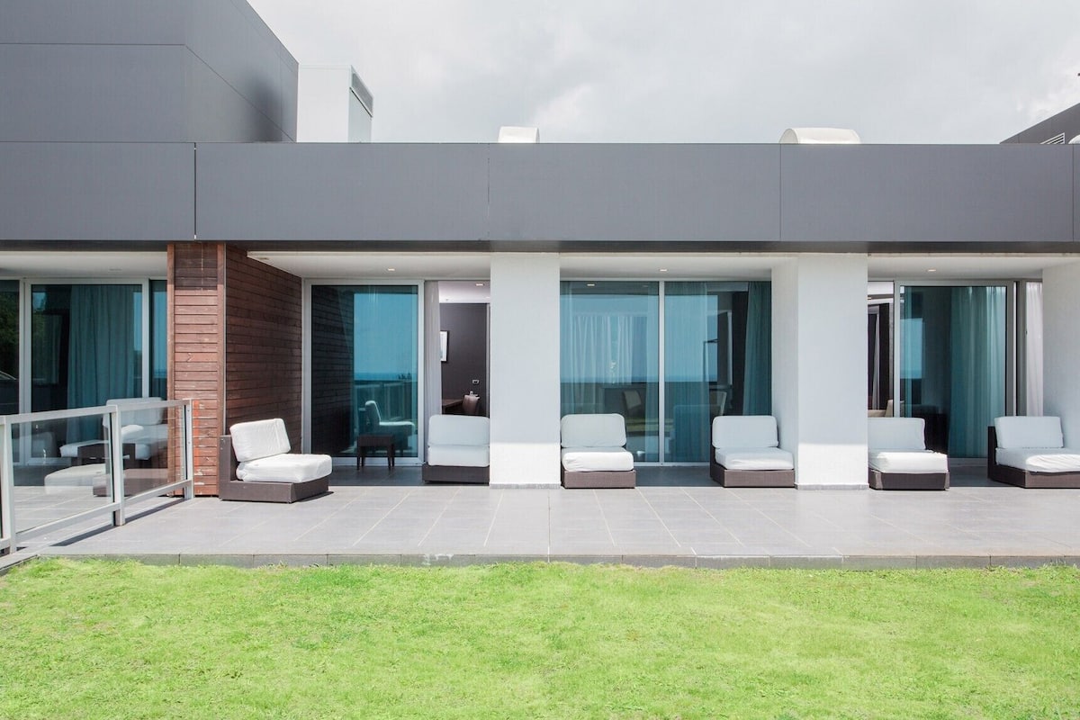 A modern hotel with a covered outdoor seating area, glass doors leading to a balcony, and a view of the sea.