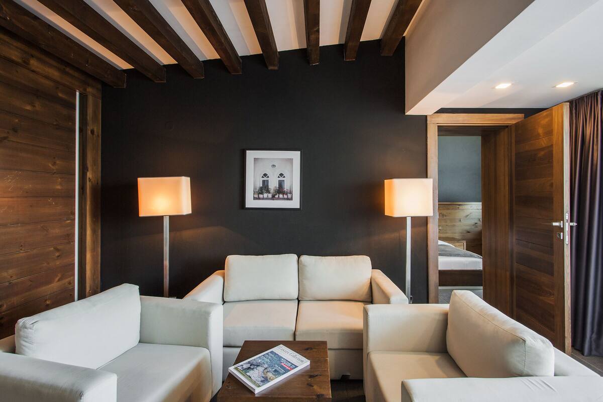 A modern hotel room with a dark accent wall, a framed picture, two bedside lamps, a white sofa, and a wooden coffee table.