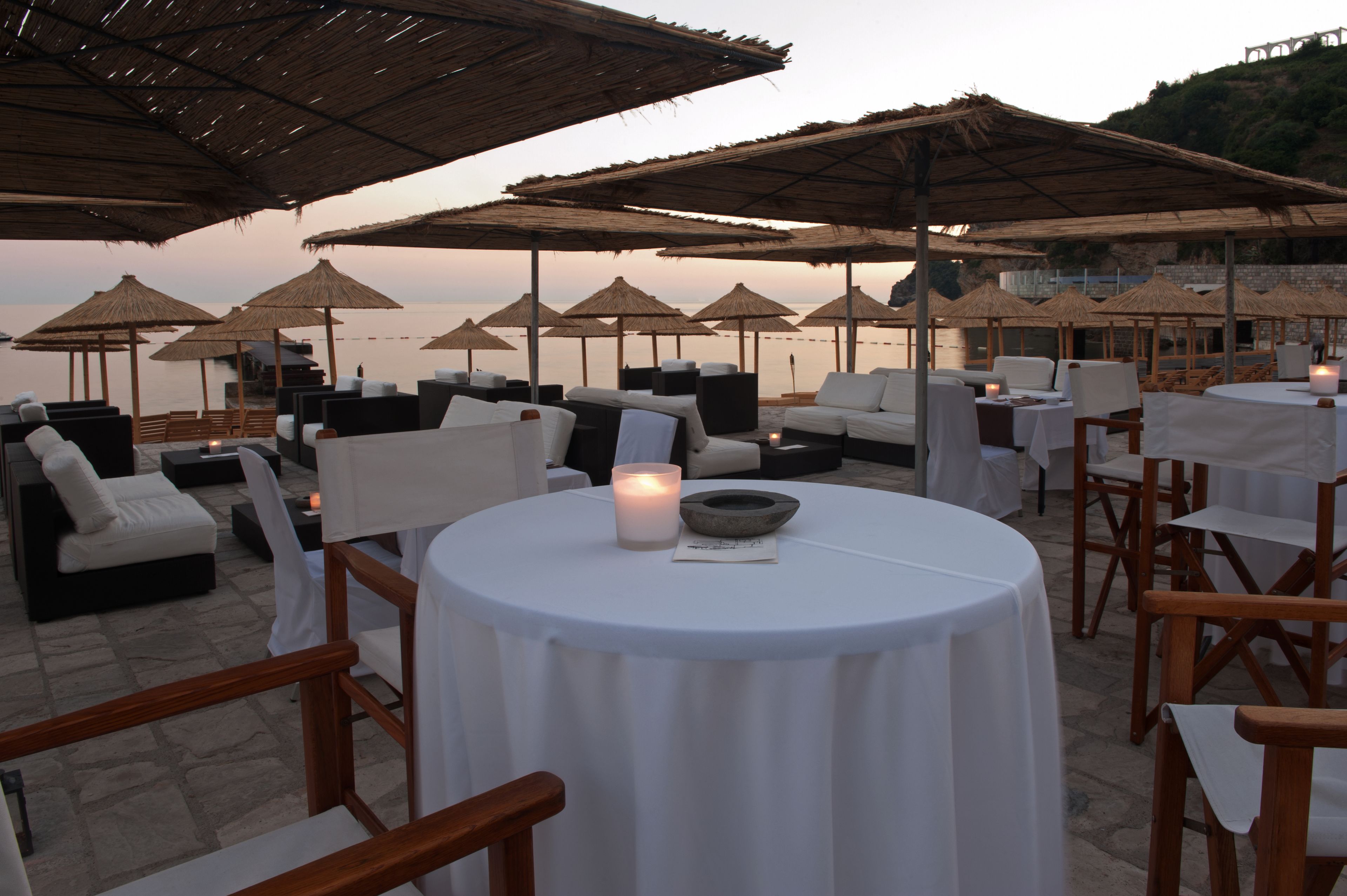 An outdoor seating area with round tables and chairs, covered by thatched umbrellas, overlooking a body of water.