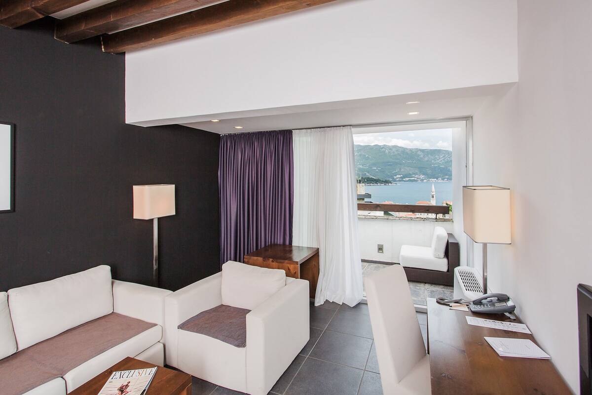 A modern hotel room with a sofa, a small table, a lamp, and a view of the sea.