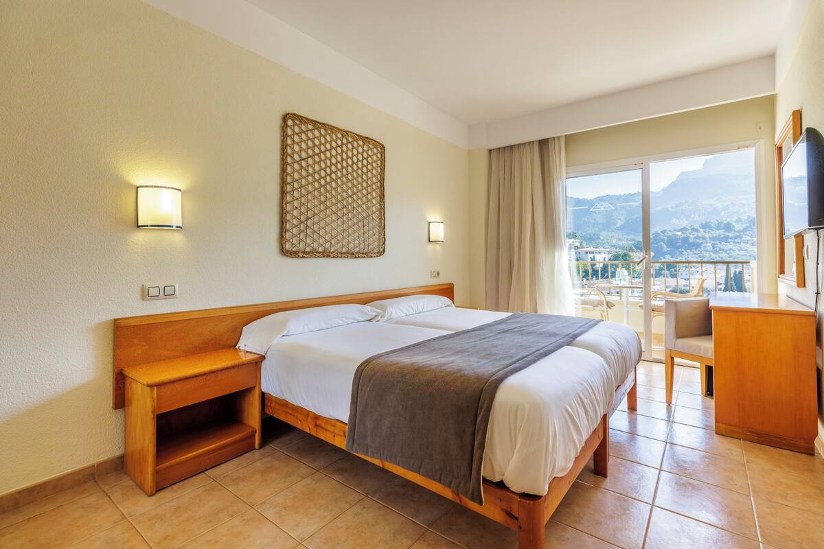 A hotel room with a large bed, a wooden nightstand, a desk, and a chair. There is a large window with a view of the mountains.