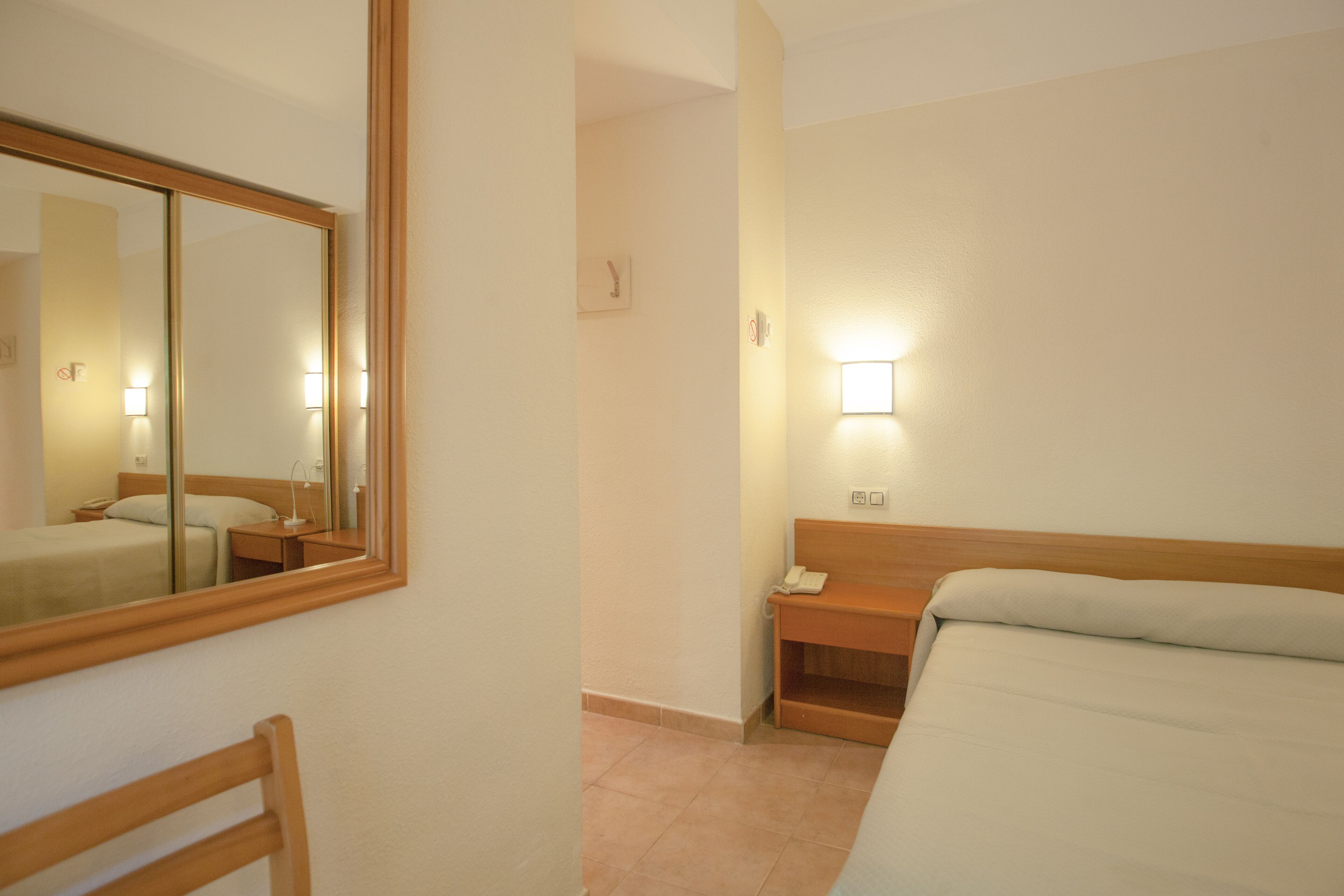 A hotel room with a bed, bedside table, mirror, and wall-mounted light.
