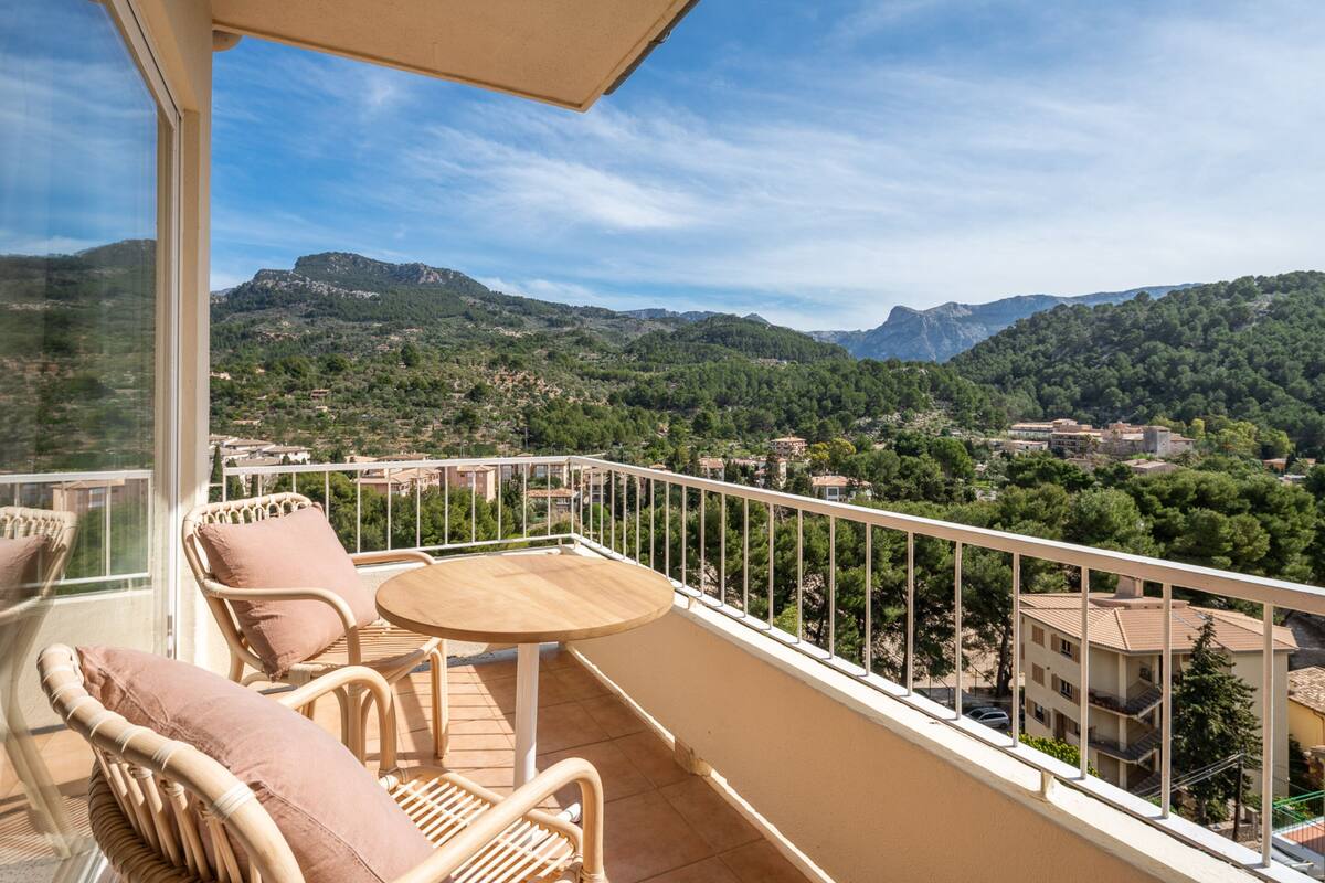 A balcony with wicker furniture and a table, overlooking a mountainous landscape with buildings in the distance.