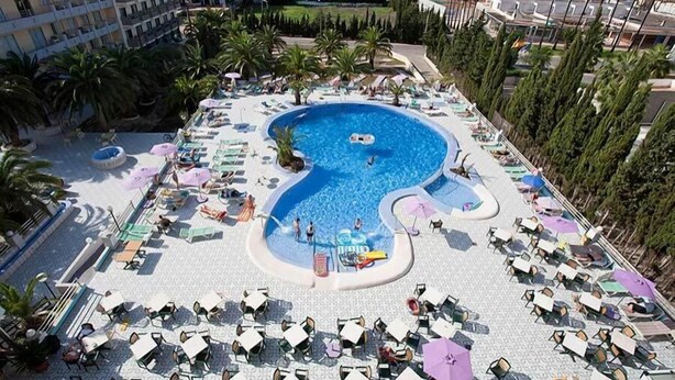 A pool area with multiple sun loungers and umbrellas, surrounded by palm trees and a building in the background. - BJ Playa Blanca (Sant Llorenc des Cardassar)