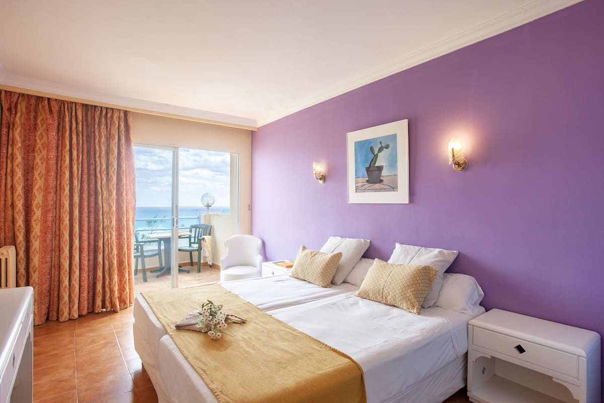 Een hotelkamer met een bed, nachtkastje, stoel, tafel en uitzicht op de oceaan door een schuifdeur.