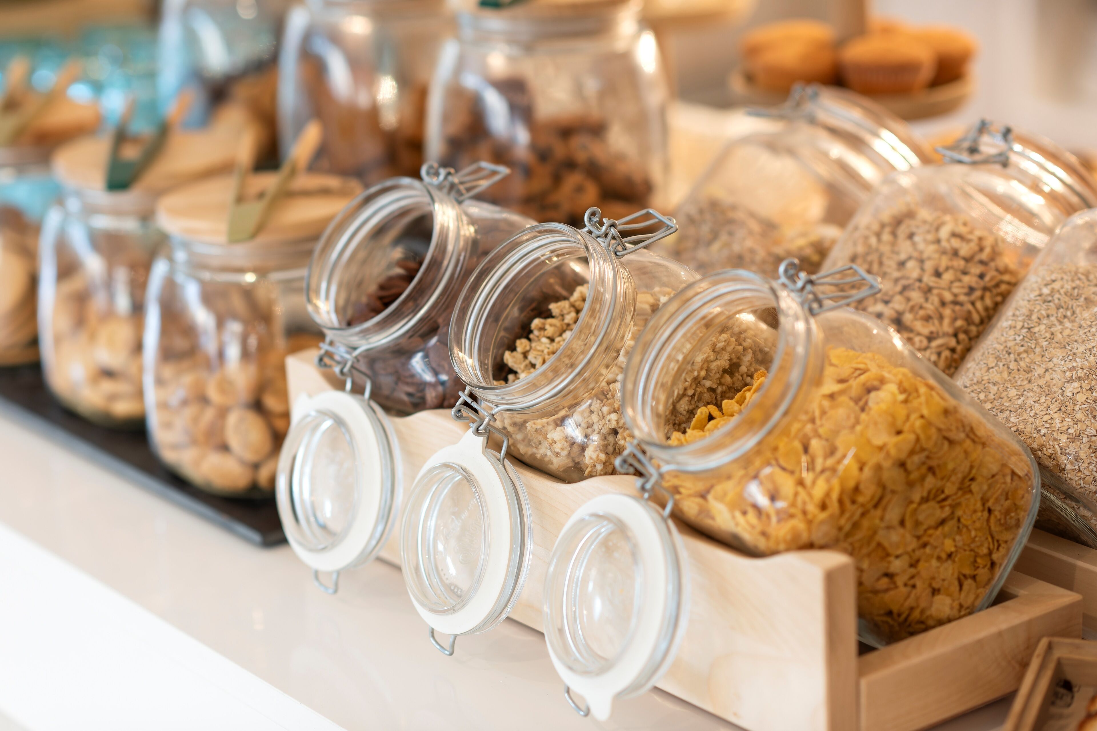 A display of various grains and seeds in glass jars with metal clasps, arranged on a wooden board.