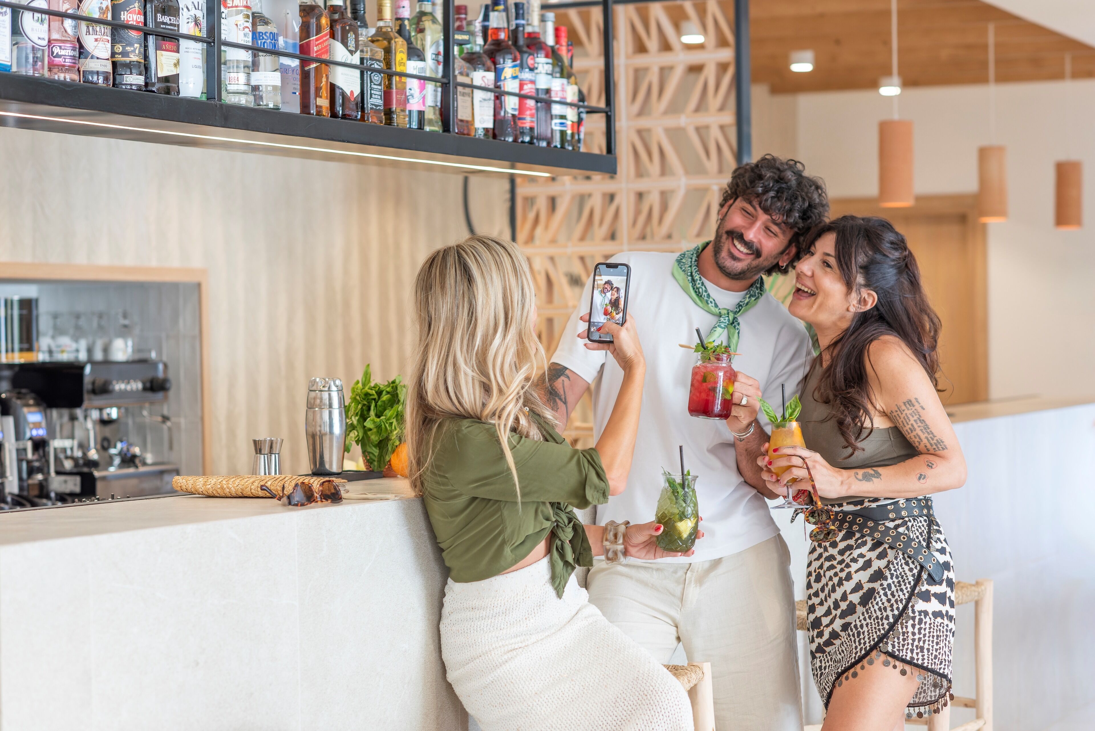 A bar area with a variety of liquor bottles on shelves, a coffee machine, and a counter with a drink and a smartphone being used for a selfie.