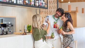 A bar area with a variety of liquor bottles on shelves, a coffee machine, and a counter with a drink and a smartphone being used for a selfie.