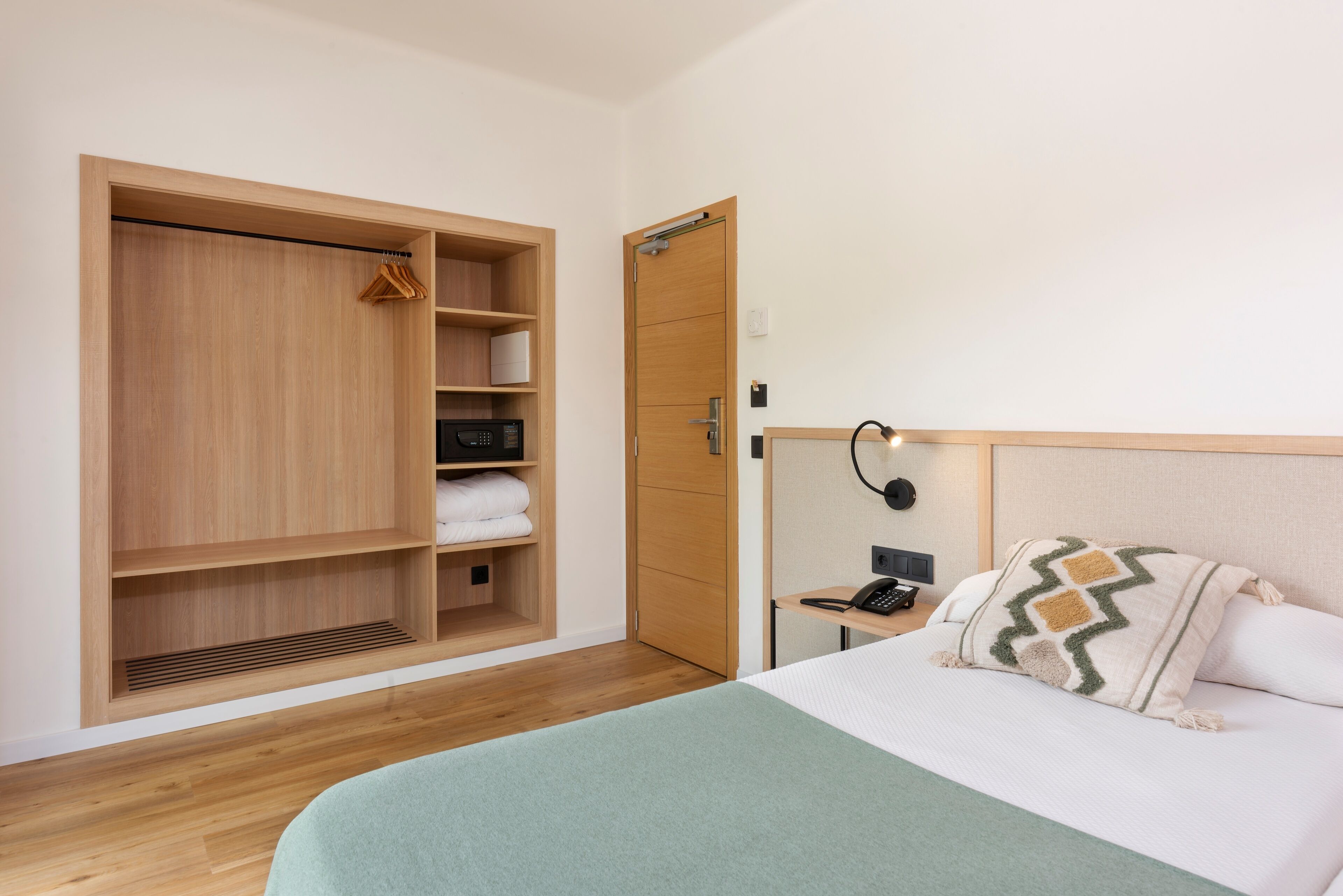 Quarto de hotel com um guarda-roupa de madeira grande, uma cama com manta verde, mesa de cabeceira com abajur e telefone.