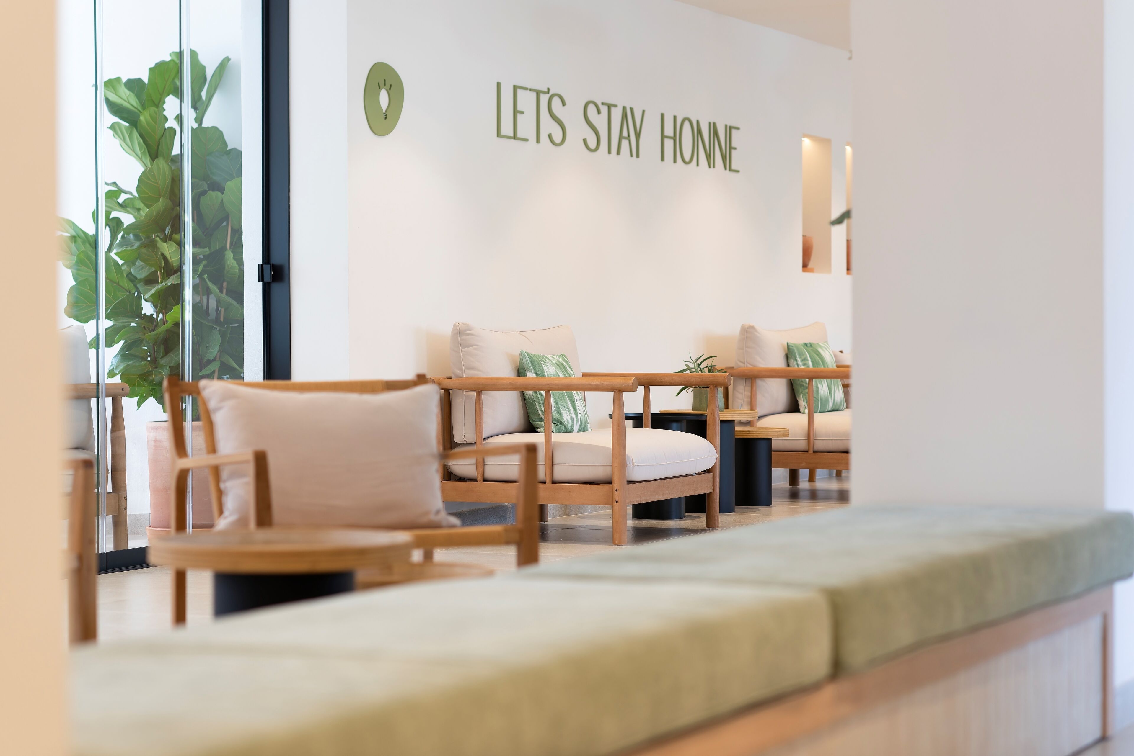 Un hall d’hôtel moderne avec un espace salon comprenant des chaises en bois et un banc. Une inscription sur le mur indique : « RESTONS À LA MAISON ».