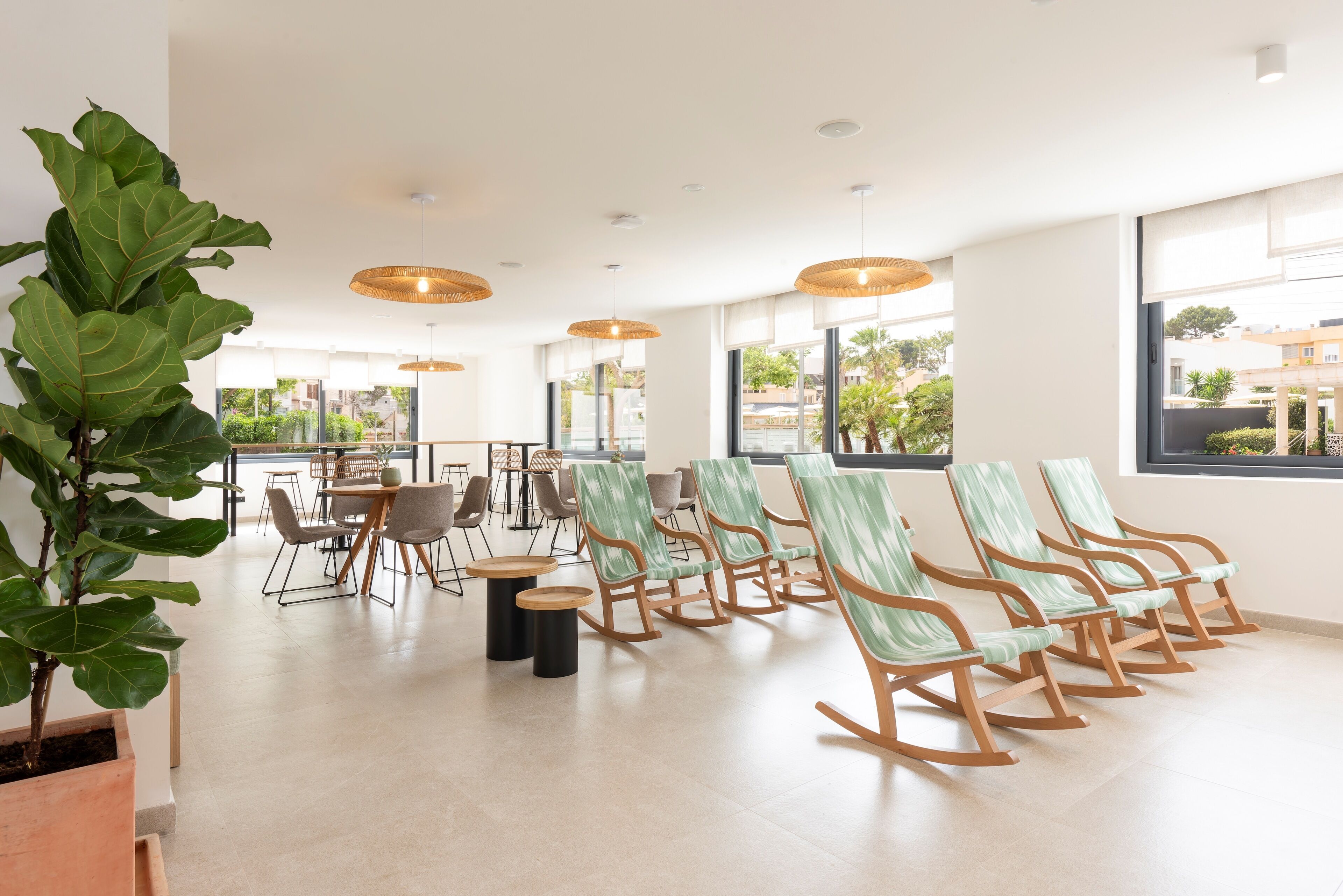 A spacious room with a long row of rocking chairs, a large potted plant, and a dining area with tables and chairs.