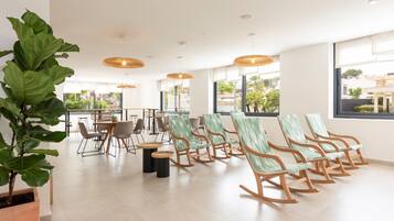 A spacious room with a long row of rocking chairs, a large potted plant, and a dining area with tables and chairs.