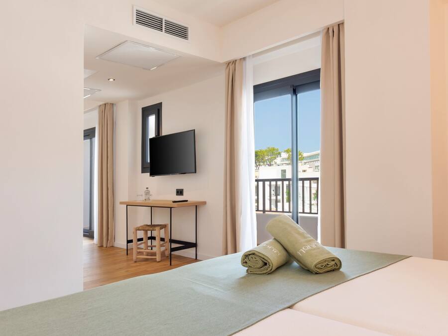 A modern hotel room with a large bed, a small desk, a television, and a view of the city through large windows.
