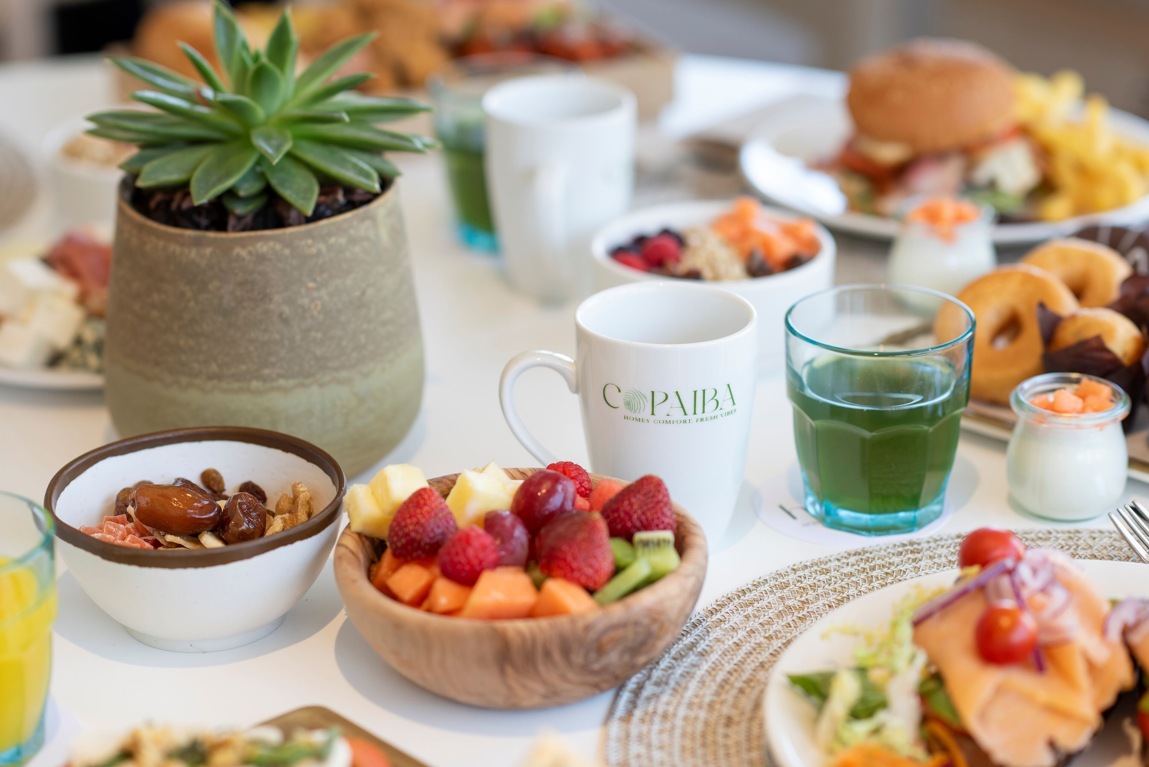 A breakfast table with various food items, including a bowl of cereal, a fruit bowl, a coffee cup, and a glass of green drink.