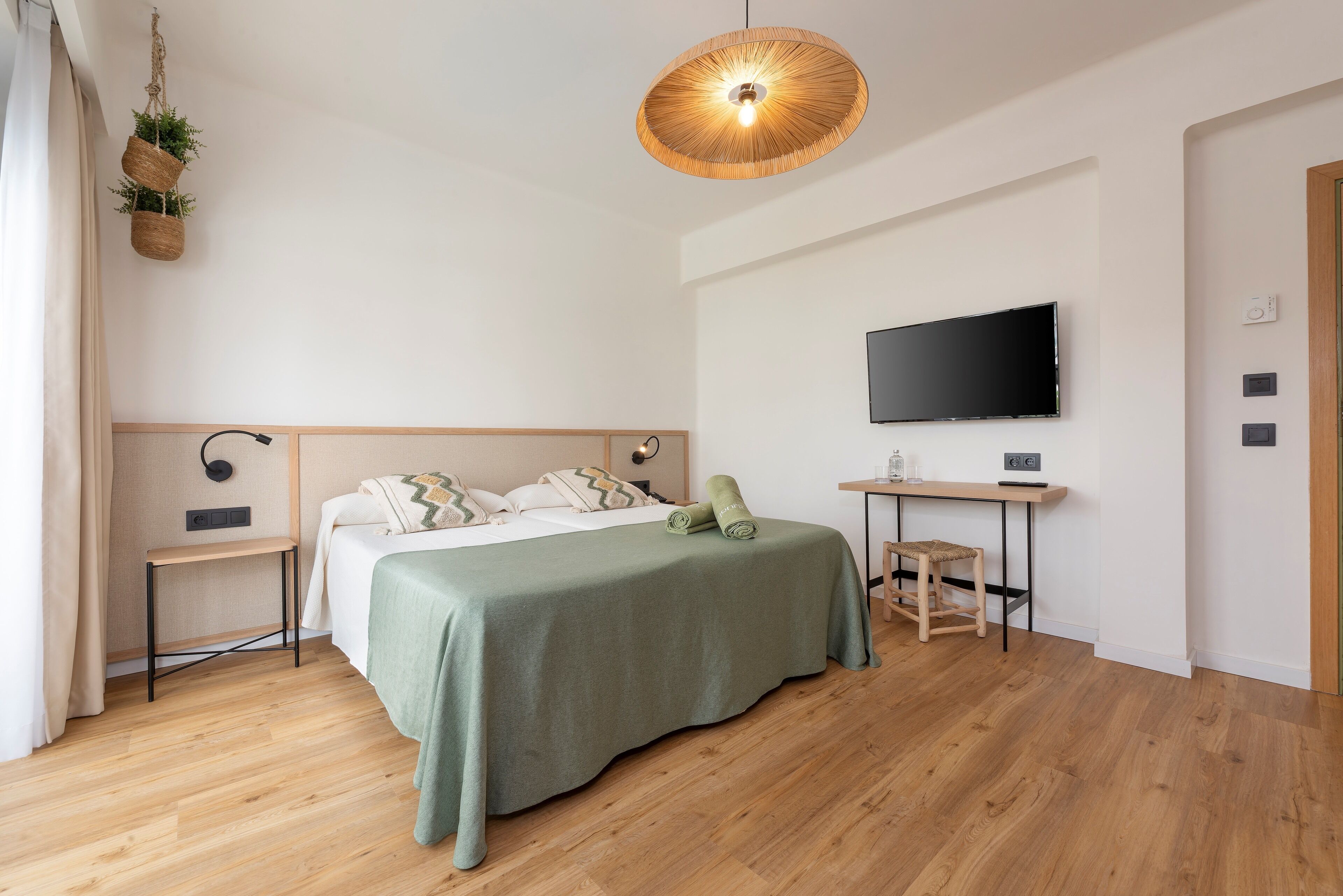 A modern bedroom with a wooden floor, a bed with a green bedspread, a bedside table, a TV mounted on the wall, and a hanging plant.