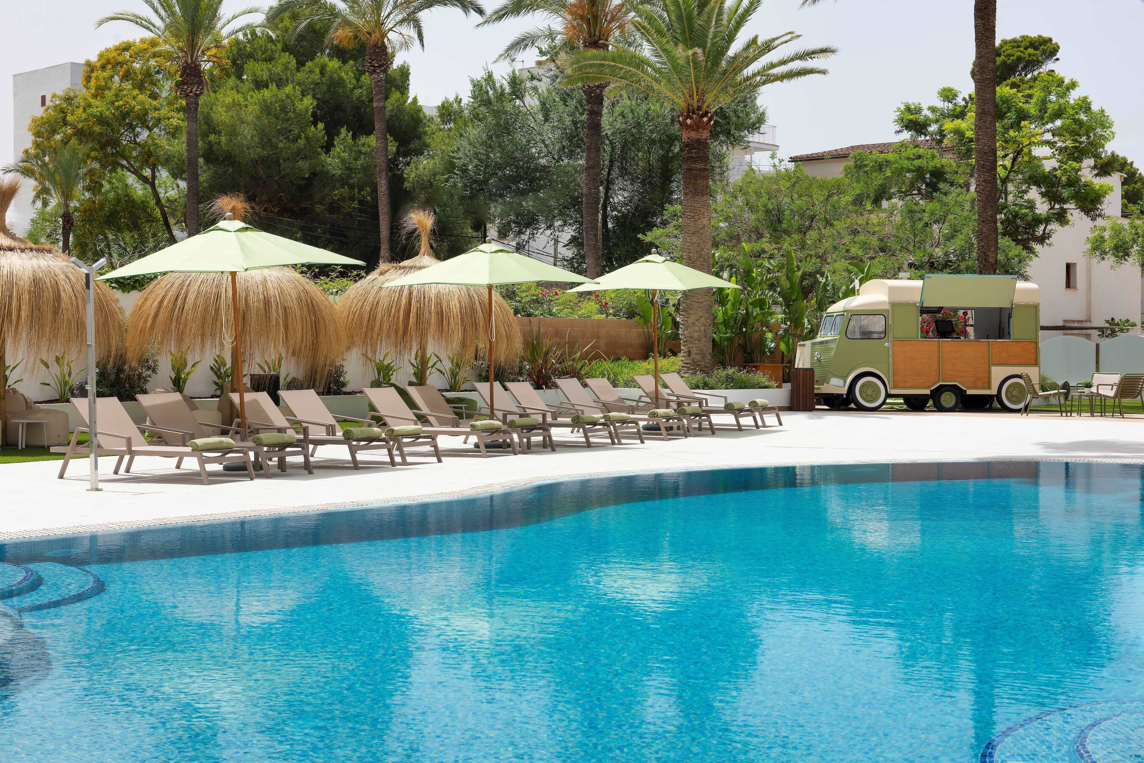 Una piscina con sillas de descanso y sombrillas, un camión de comida y palmeras.
