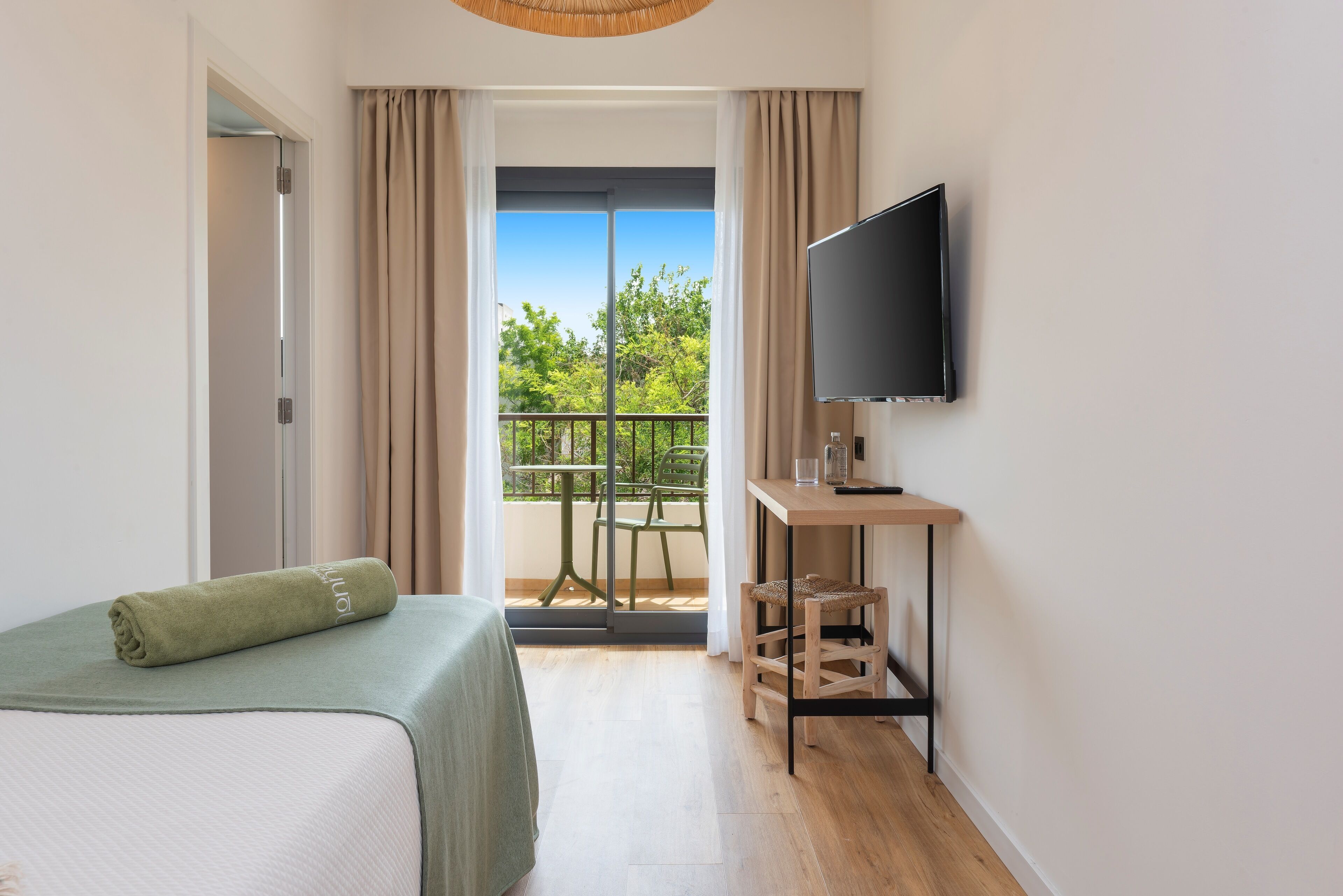 Habitación de hotel con cama, televisión, una mesita y balcón con vistas a árboles.