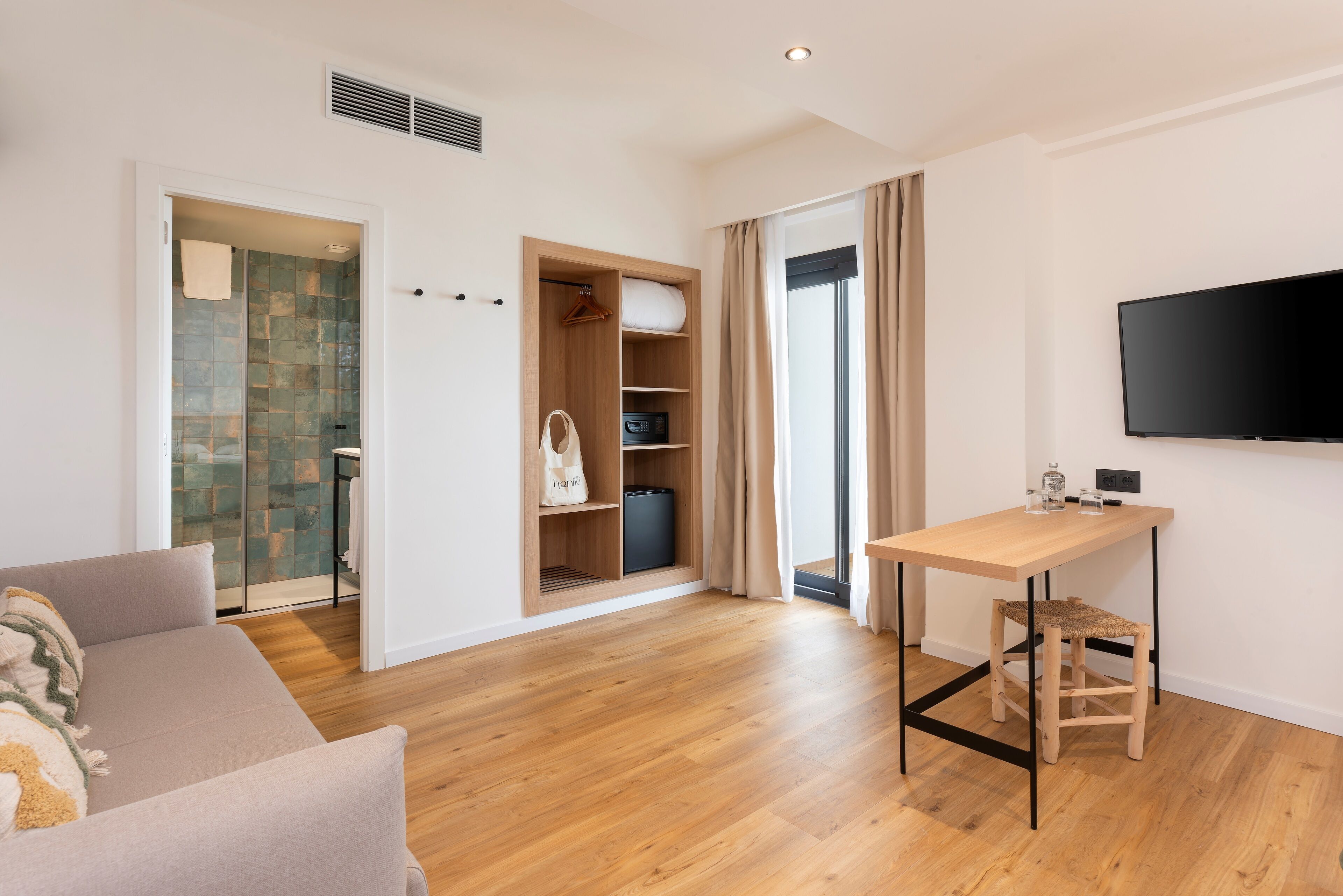 A modern hotel room with a wooden floor, a sofa, a desk with a chair, a TV mounted on the wall, and a walk-in closet with a hanging bag.