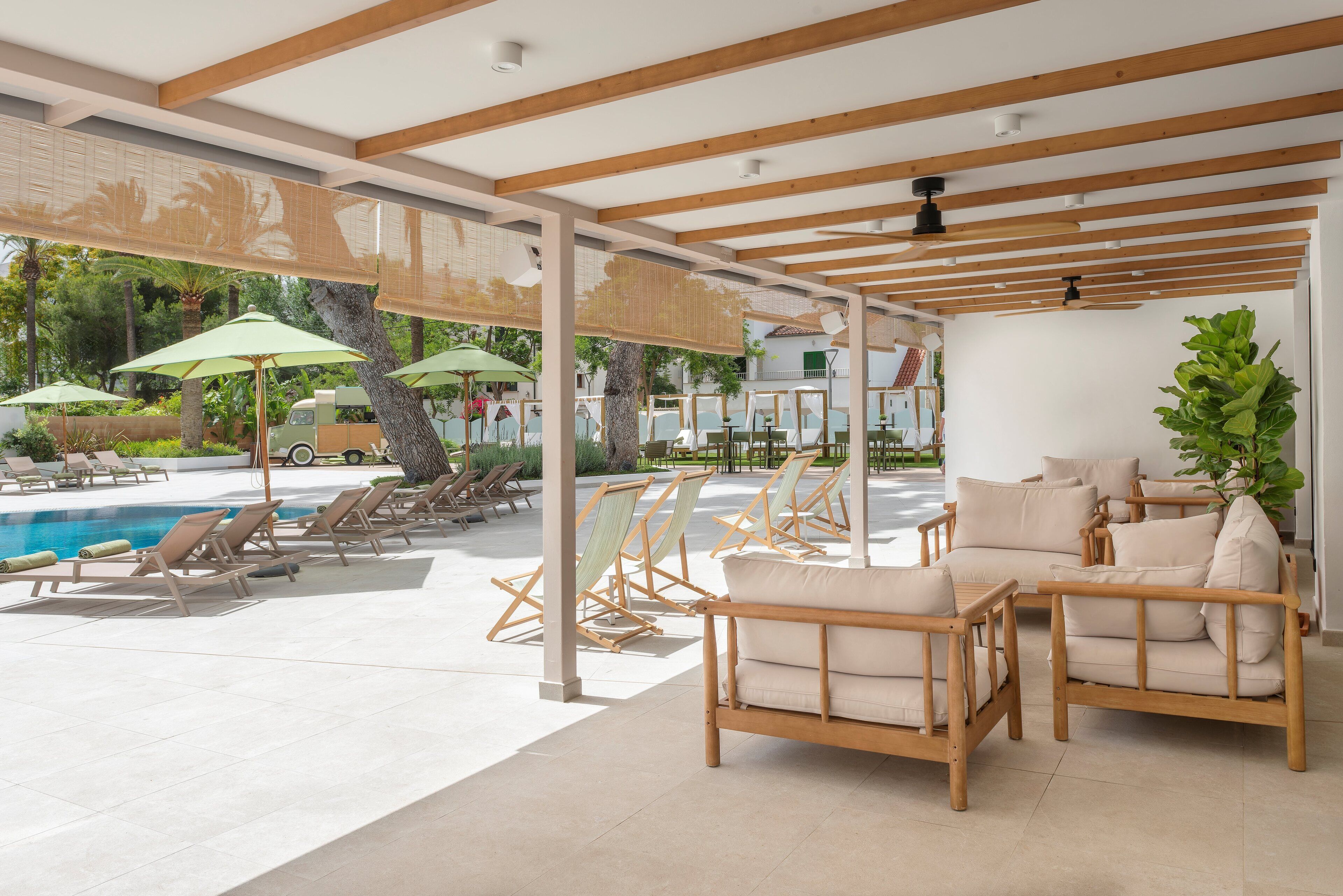 Un área junto a la piscina con sillones, una pérgola de madera y un espacio de descanso con cojines.