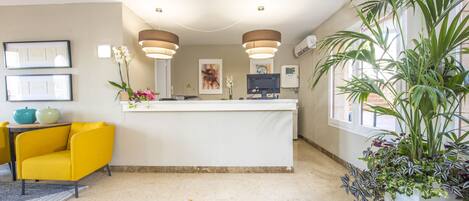 A reception area with a yellow armchair, a reception desk, pendant lighting, framed artwork, and potted plants.