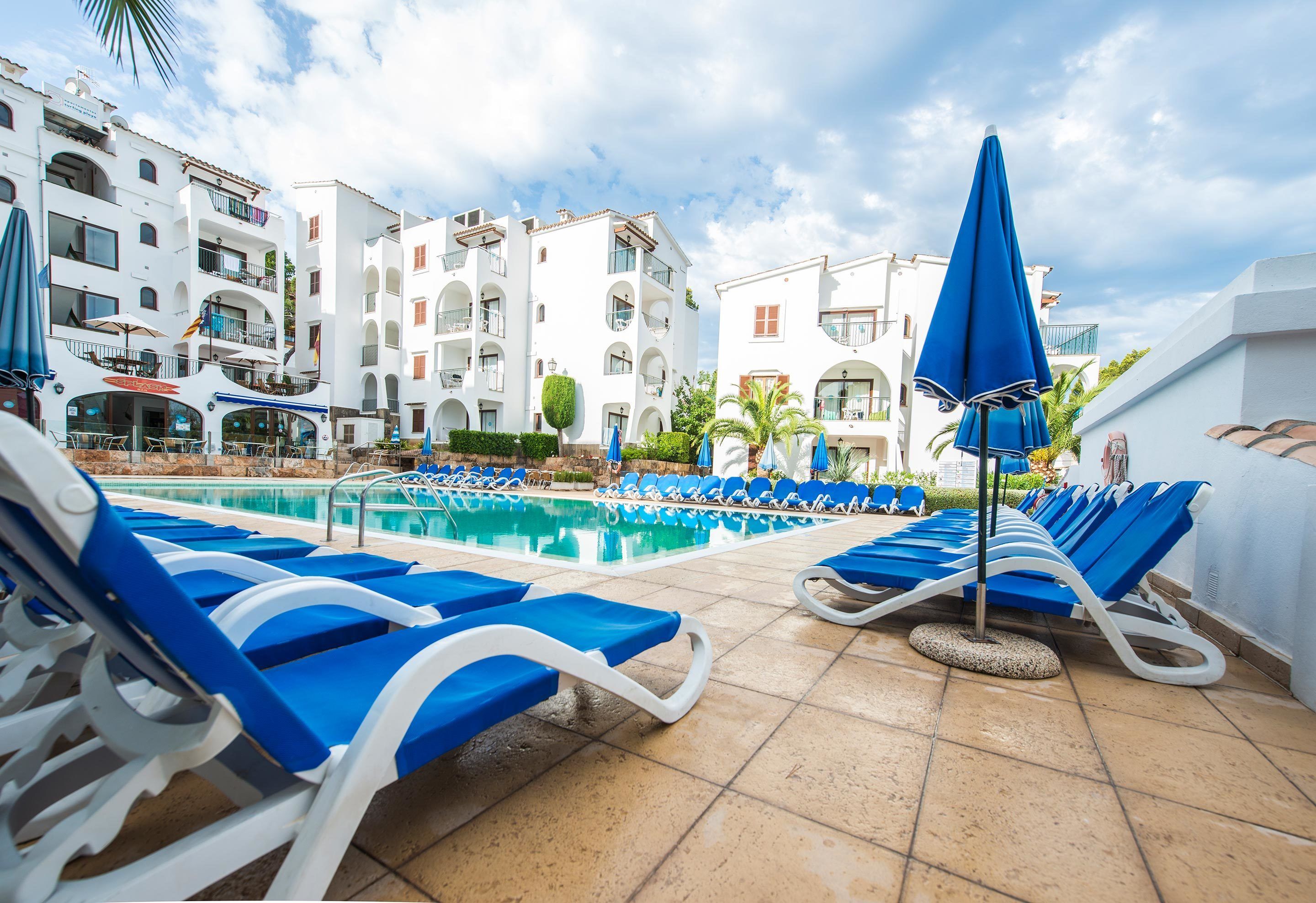 A poolside area with blue lounge chairs and umbrellas, surrounded by white buildings with balconies and a clear sky.