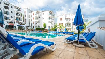 A poolside area with blue lounge chairs and umbrellas, surrounded by white buildings with balconies and a clear sky.